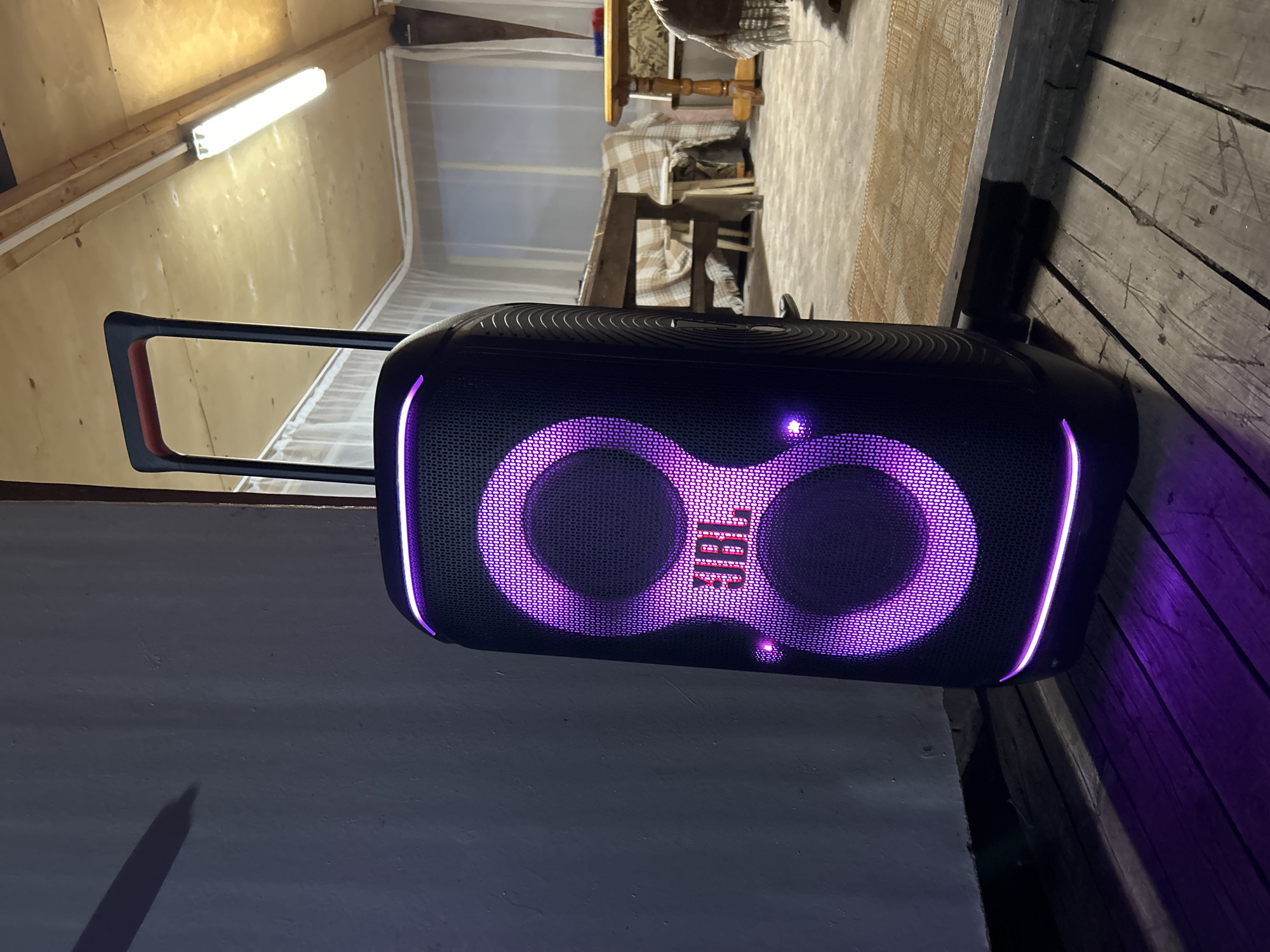 A large, black portable speaker with vibrant purple LED lights illuminating its circular speaker grilles and outlines stands prominently in what appears to be a rustic indoor or semi-enclosed space. The speaker features the brand name "JBL" in red-pink lettering on its front face and has a retractable handle, partially visible at the top left.
The setting suggests a practical or informal area, possibly a workshop, a veranda, or a room in a cabin or dacha, consistent with the location context of Foki, Russia. To the top left, a plain wooden wall or ceiling is visible, featuring a long, bright fluorescent light fixture attached to a wooden beam. Further back and to the right, a simple wooden table covered with a light-colored checkered cloth and a chair with a blanket are visible, indicating a functional living or dining area. The floor beneath the table appears to be a light-colored mat or rug.
In the foreground to the bottom left, there's a plain, darker wall or partition, casting a faint shadow from an unseen object. To the right of the speaker, a dark wooden floor or decking with visible planks and gaps stretches into the background. The overall ambiance is dim, relying on the artificial glow from the speaker's lights and the overhead fluorescent fixture, suggesting the photograph was taken in the evening or at night. The speaker is actively on, with its lights creating a festive or atmospheric effect, implying music might be playing or an event is taking place.