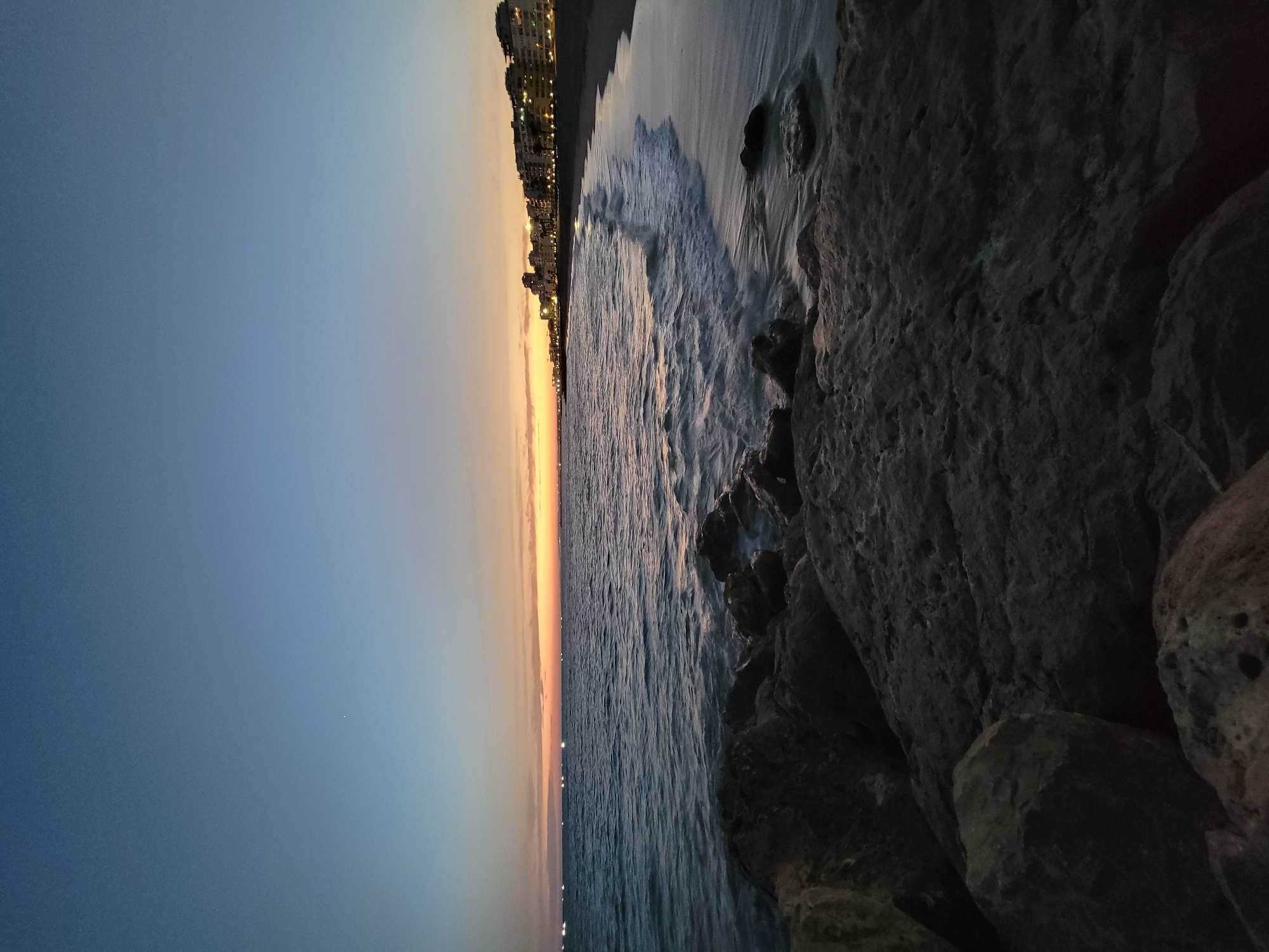 The image shows a twilight view of the beach in Quarteira, Portugal. The sky is a gradient of dark blue at the top, transitioning to lighter blues and then a warm orange and yellow band along the horizon, indicating sunset or sunrise. The sea is calm with gentle waves washing onto the sandy beach. In the foreground, there are large, dark, rough-textured rocks and boulders. Along the coastline in the distance, a line of buildings, likely hotels and apartments, is visible, with lights beginning to illuminate them. Several small lights, possibly from boats, are scattered across the sea. The overall scene is serene and captures the transition from day to night.