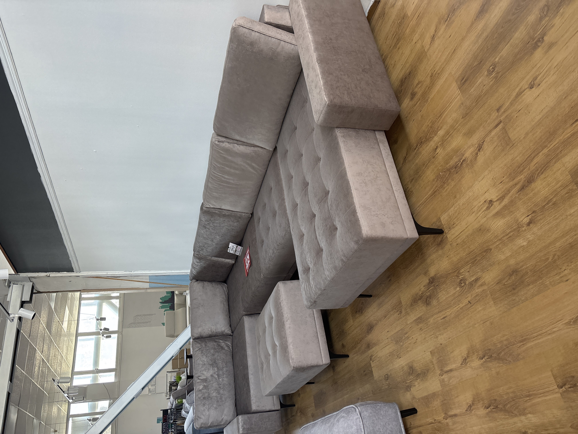 A large, L-shaped sectional sofa with tufted seating and taupe upholstery is displayed in a showroom. The sofa is composed of multiple modular sections, including a corner piece and a chaise lounge. Two square ottomans with matching upholstery and dark, tapered legs are placed in front of the sectional. The flooring is a light brown wood-effect laminate. In the background, a white wall is visible, and the ceiling features industrial lighting and structural elements. The overall setting appears to be a furniture store, likely within the city of Zaporizhzhia, Ukraine, given the provided context.