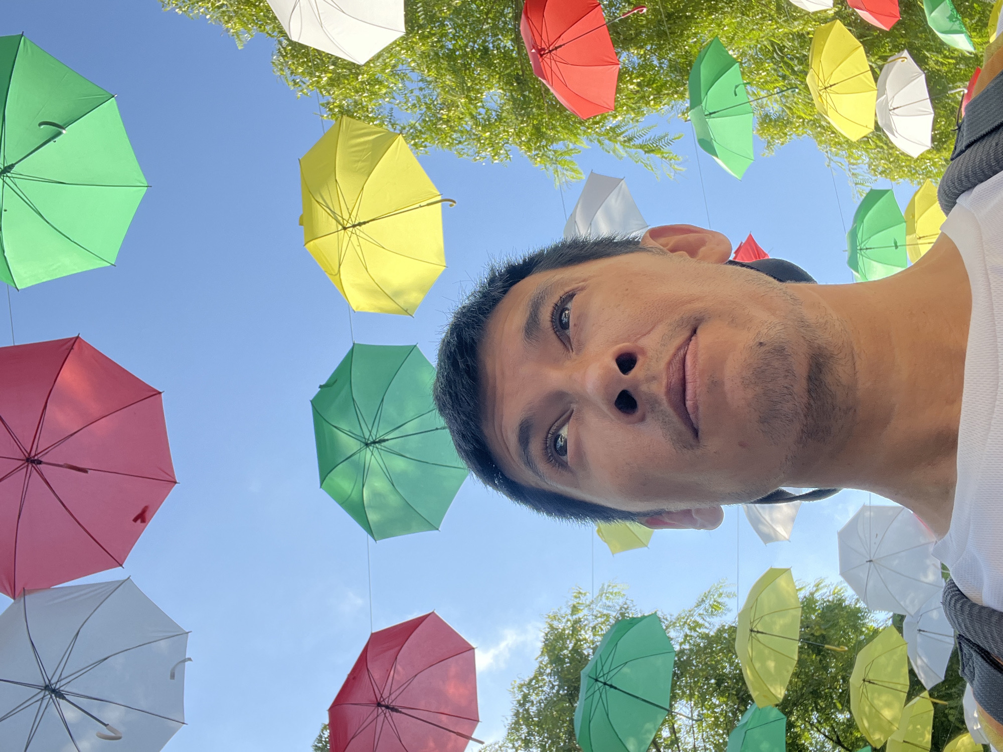 The image shows a low-angle, wide shot of a man's face and upper chest, looking upwards. He has dark hair and stubble. Above him, a clear blue sky is filled with numerous colorful umbrellas of various sizes and colors, including white, pink, green, and yellow, suspended on wires. Some green foliage from trees is visible on the right side of the image, with branches reaching into the frame. The scene appears to be outdoors, potentially a street or public area decorated with the umbrellas. The overall impression is a vibrant, summery atmosphere. This scene is from the Rambla Nova in the city of Tarragona, Spain, a popular pedestrian street known for its art installations and decorations during certain times of the year, including the display of colorful umbrellas.