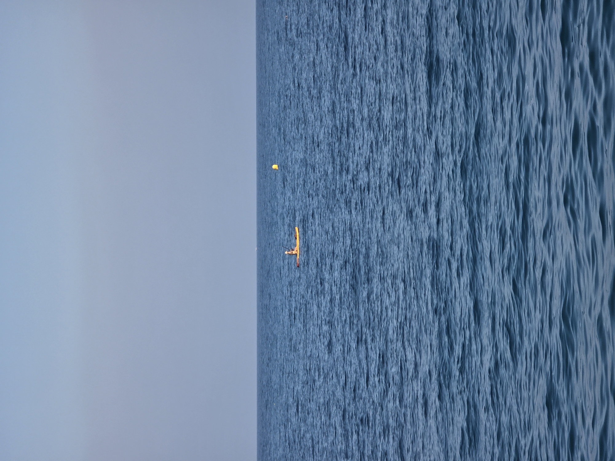 A lone individual wearing a hat is seated on a yellow paddleboard in the deep blue water of the Mediterranean Sea. The paddleboarder is positioned in the middle of the frame, approximately one-third of the way from the left edge, and appears to be paddling. The water is calm with gentle ripples. In the distance, to the right of the paddleboarder, a yellow buoy is visible. The horizon line is clearly defined, separating the sea from a pale blue sky that is mostly clear, with a faint haze. The scene suggests a tranquil day on the water, likely off the coast of Nice, France.
