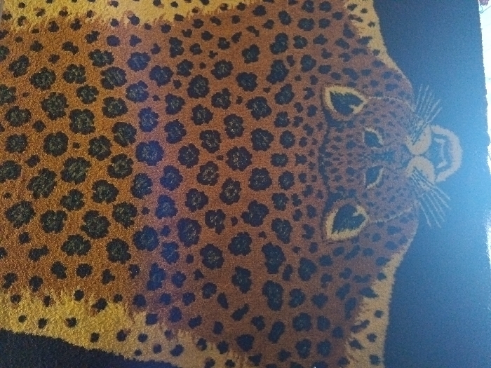 A leopard-print rug or carpet dominates the view. The design features dense groupings of irregular black spots over a tan or brownish backdrop. The head of the leopard is positioned on the right side, featuring a symmetrical design with pointed ears and whiskers. The rug appears to be placed on a darker colored surface, creating a contrasting background.