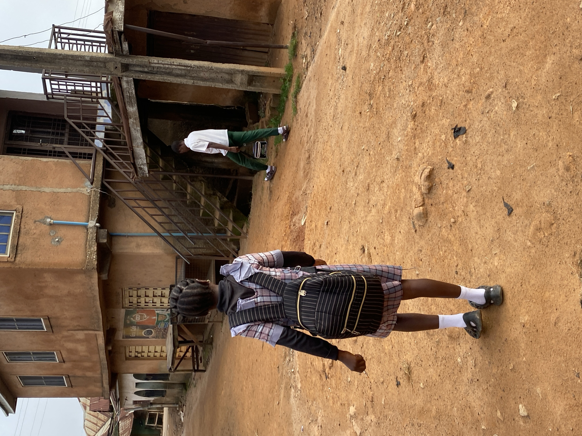The image depicts a dusty, unpaved street scene in Jos, Nigeria, with a multi-story building constructed of earth-toned stucco prominently featured. In the foreground, a young Black girl wearing a plaid dress, black long-sleeved shirt, white socks, and dark clogs walks away from the camera, carrying a striped backpack. Further down the street, another Black individual, possibly a boy, walks away from the camera wearing a white short-sleeved shirt and green pants, carrying a black and white bag. The building has multiple windows, an external staircase with metal railings, and doorways. Several corrugated metal roofs are visible in the background, indicative of residential structures. The ground is predominantly dirt and sand, with scattered debris. The overall impression is a common urban or semi-urban setting in Jos.