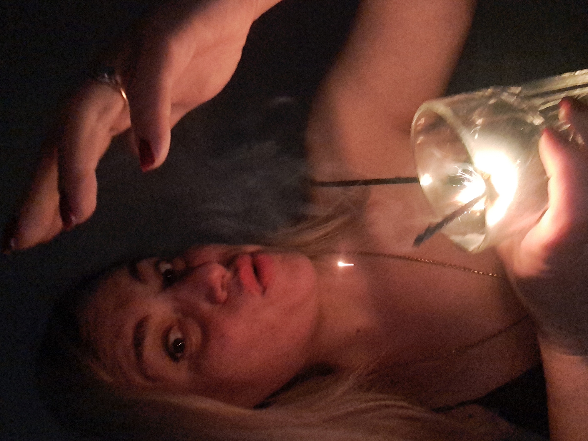 A woman lies on her back in a dimly lit environment. She is fair-skinned with blonde hair. She holds a glass container with a sparkler inside, which is emitting light and smoke. Her other hand is raised, seemingly hovering above her. She wears a thin gold necklace and a ring on one of her fingers. It is impossible to determine the location of Elektrostal, Russia from this image.