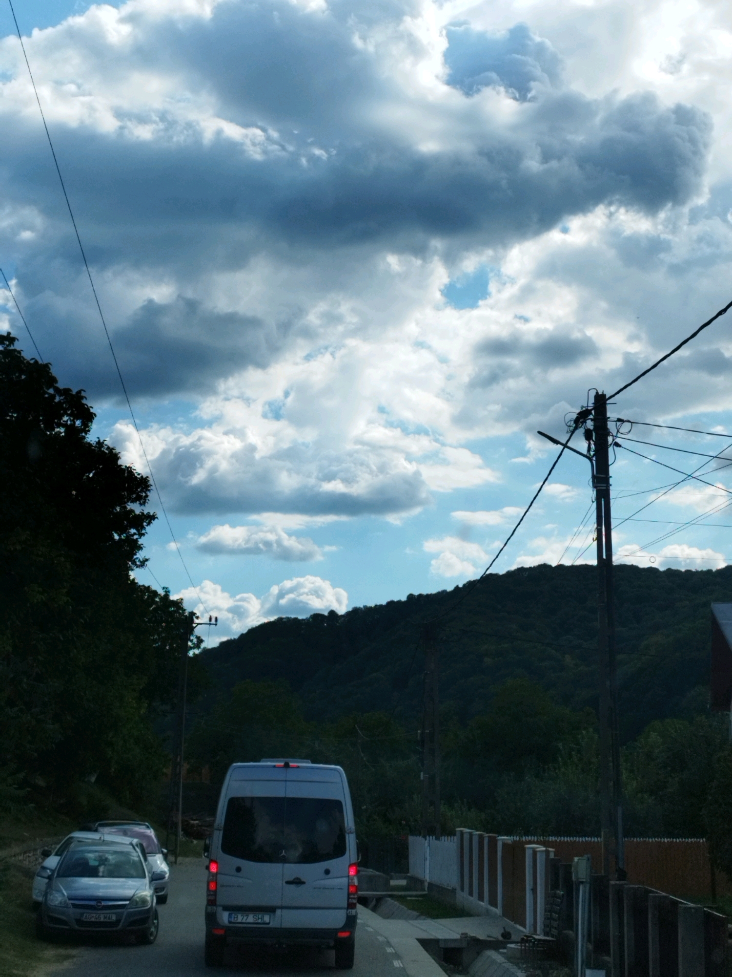 The image captures a scene on a road in the vicinity of Oeștii Ungureni, Romania. A white van is driving away from the camera, its rear visible. To the left of the van, several cars are parked on the side of the road. The road is flanked by trees and a lush, green hillside covered in dense forest rises in the background. A utility pole with multiple wires is prominent on the right side of the road, silhouetted against a dramatic sky filled with both dark and bright clouds. The overall impression is of a rural or semi-rural setting under a dynamic, overcast sky.