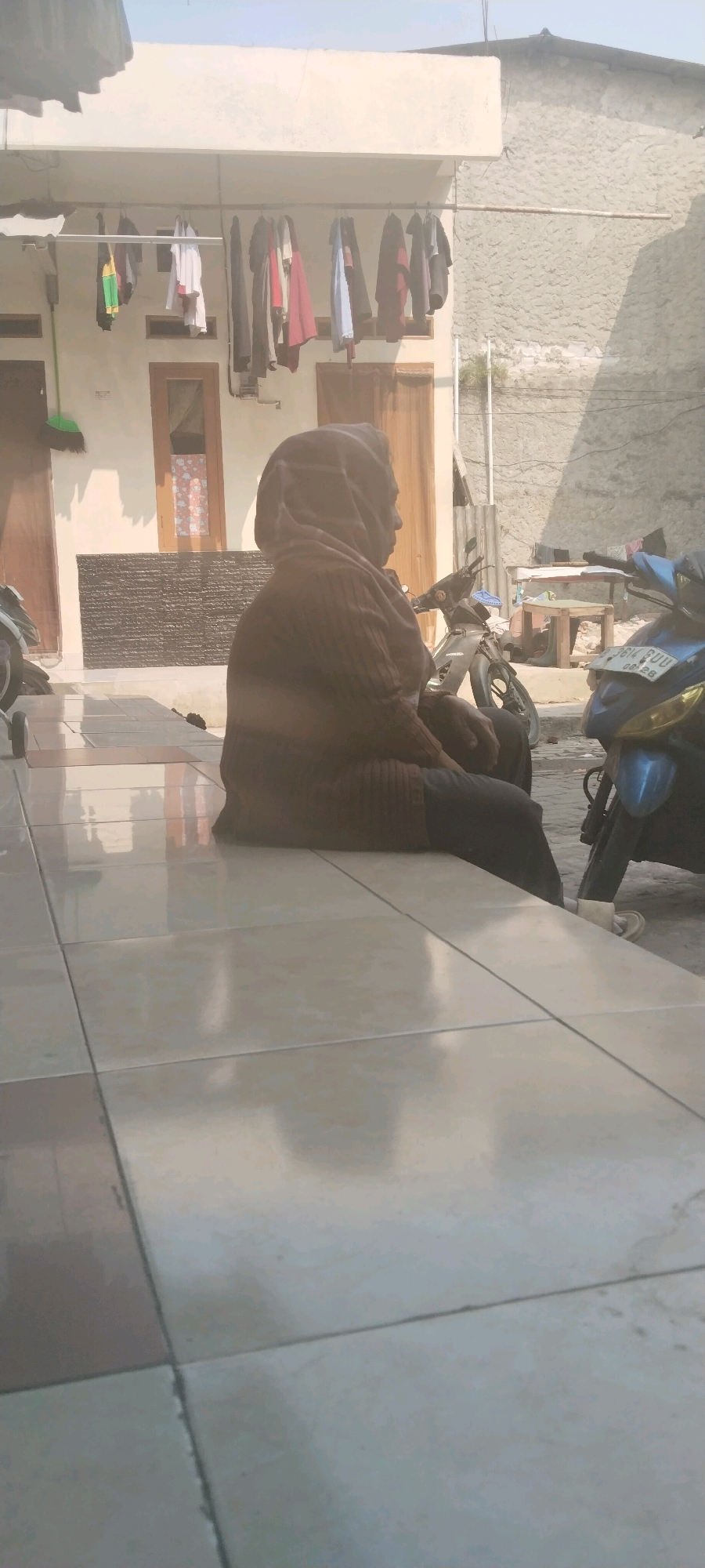 The image shows a person wearing a brown headscarf and a patterned brown sweater, sitting on a tiled floor. The person is facing to the right, away from the camera. In the background, there is a building with clothes hanging on a clothesline. Several motorcycles are parked in front of the building. The environment appears to be an outdoor area, possibly a courtyard or alleyway, in a residential setting. The overall lighting suggests it is daytime, with sunlight casting shadows. There are no discernible elements that definitively place the scene within West Jakarta, Indonesia; however, the architectural style and presence of motorcycles are common in such urban environments.