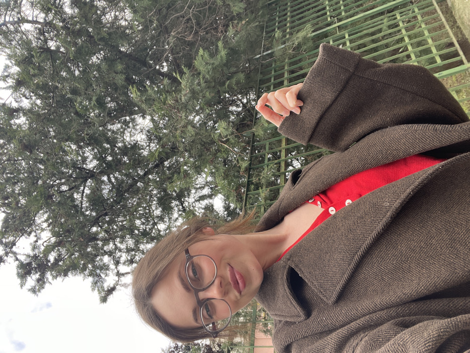 A female subject is depicted in an outdoor self-portrait in Cava de' Tirreni, Italy, under an overcast sky. She occupies the lower half of the frame, facing the camera with a subtle smile. She wears round, dark-framed eyeglasses, a dark brown herringbone-patterned overcoat with wide lapels, and a red v-neck top with white buttons. Her right hand holds the cuff of her left sleeve. Behind her, a large evergreen tree with dense, dark green foliage and visible branches rises against the sky. A dark green metal fence is visible in the background, extending from the center to the right, and a light pink building wall is partially visible on the far left. The precise location within Cava de' Tirreni cannot be determined from the visual evidence.