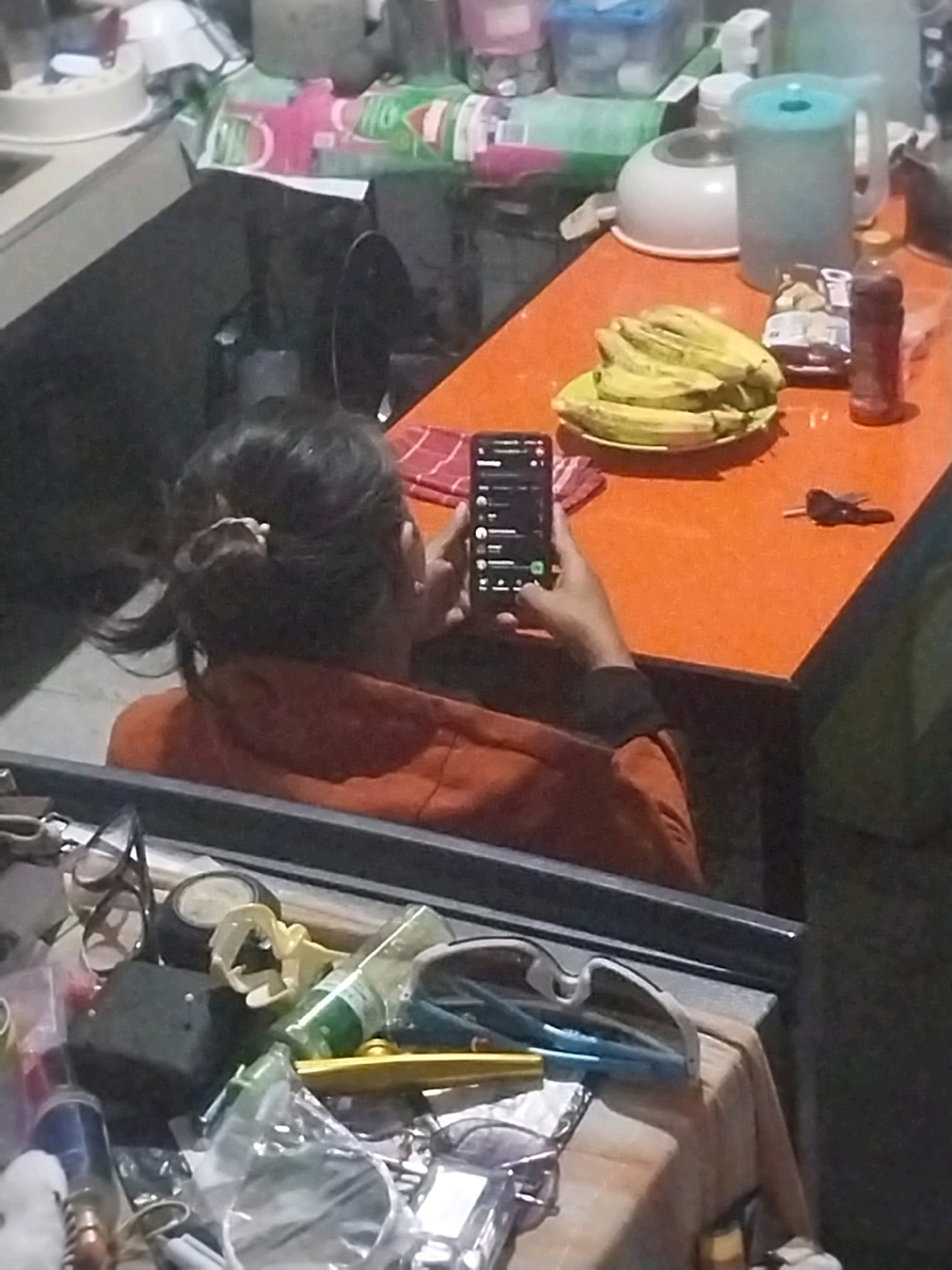 The image depicts an indoor scene, likely a kitchen or dining area, within a residential setting in Blitar, Indonesia. A woman with dark hair, pulled back in a ponytail, is seated at an orange table, looking at her smartphone. She is wearing an orange long-sleeved shirt. On the table, a plate of bananas is visible, along with a bottle of liquid and a small packet of snacks. A red and white checkered cloth is placed near the bananas. In the foreground, a reflection in a mirror or glass surface reveals a collection of various objects, including eyeglasses, a black rectangular object, a green bottle, a yellow object, and other miscellaneous items. The overall environment appears to be somewhat cluttered, suggesting a lived-in space. The lighting suggests it is either evening or indoors with artificial light.