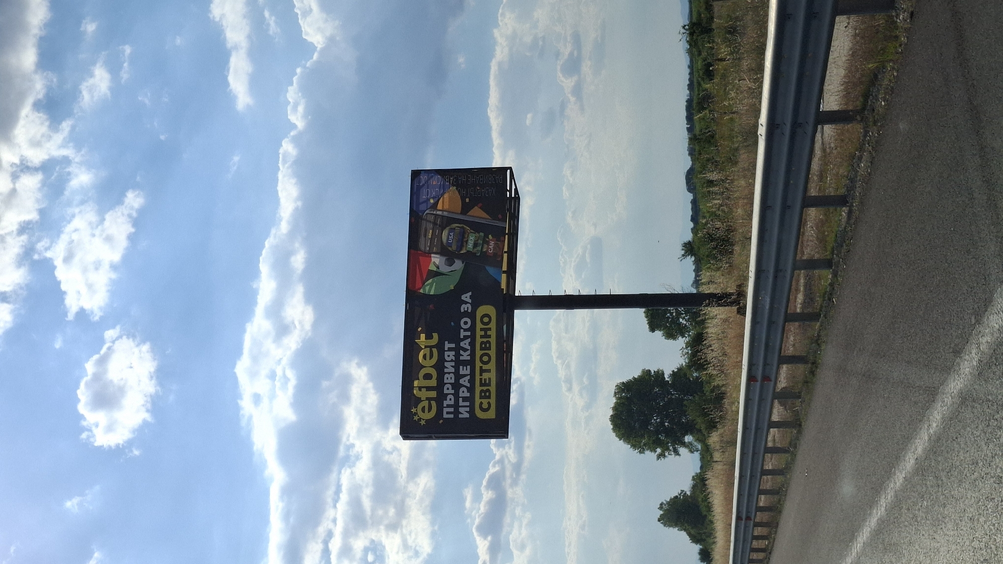 The image shows a billboard with the Efbet logo and text in Bulgarian. The billboard is located in Malo Konare, Bulgaria, alongside a highway. The billboard advertises Efbet, a betting company. The text on the billboard reads "първият играе като за световно", which translates to "the first plays like for the world". The billboard features an image of a mobile phone displaying betting options for different countries. The background consists of a blue sky with white clouds, trees, and fields. A guardrail is visible in the foreground, separating the highway from the roadside. The lighting suggests it is daytime, possibly late afternoon due to the direction of shadows. No people are visible in the image.