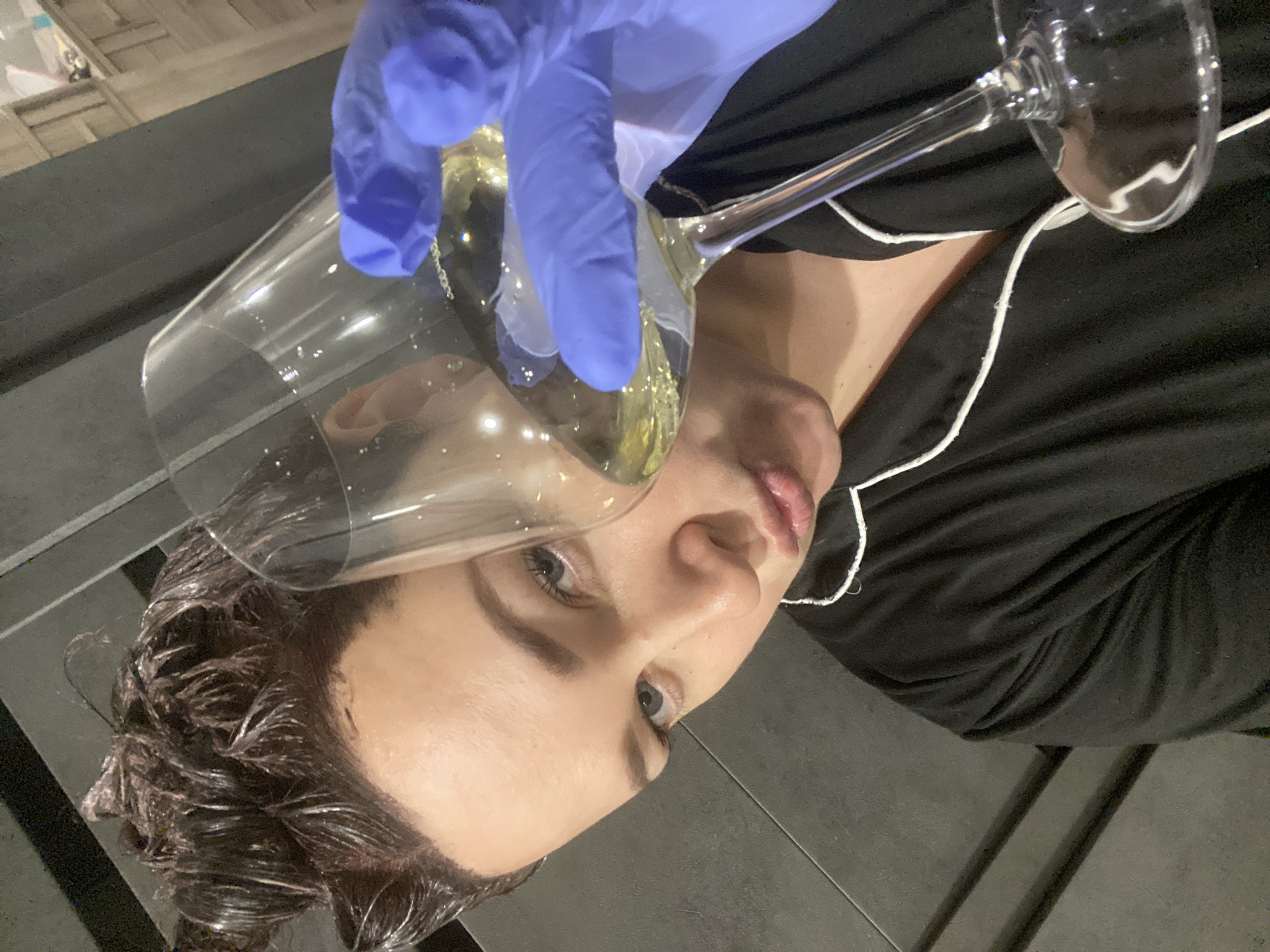 A woman wearing a black top with white trim is having a clear liquid poured from a clear glass into her hair. The hand pouring the liquid is wearing a blue glove. The woman has short brown hair and is facing the camera with her head tilted. The location is not discernible from the image.