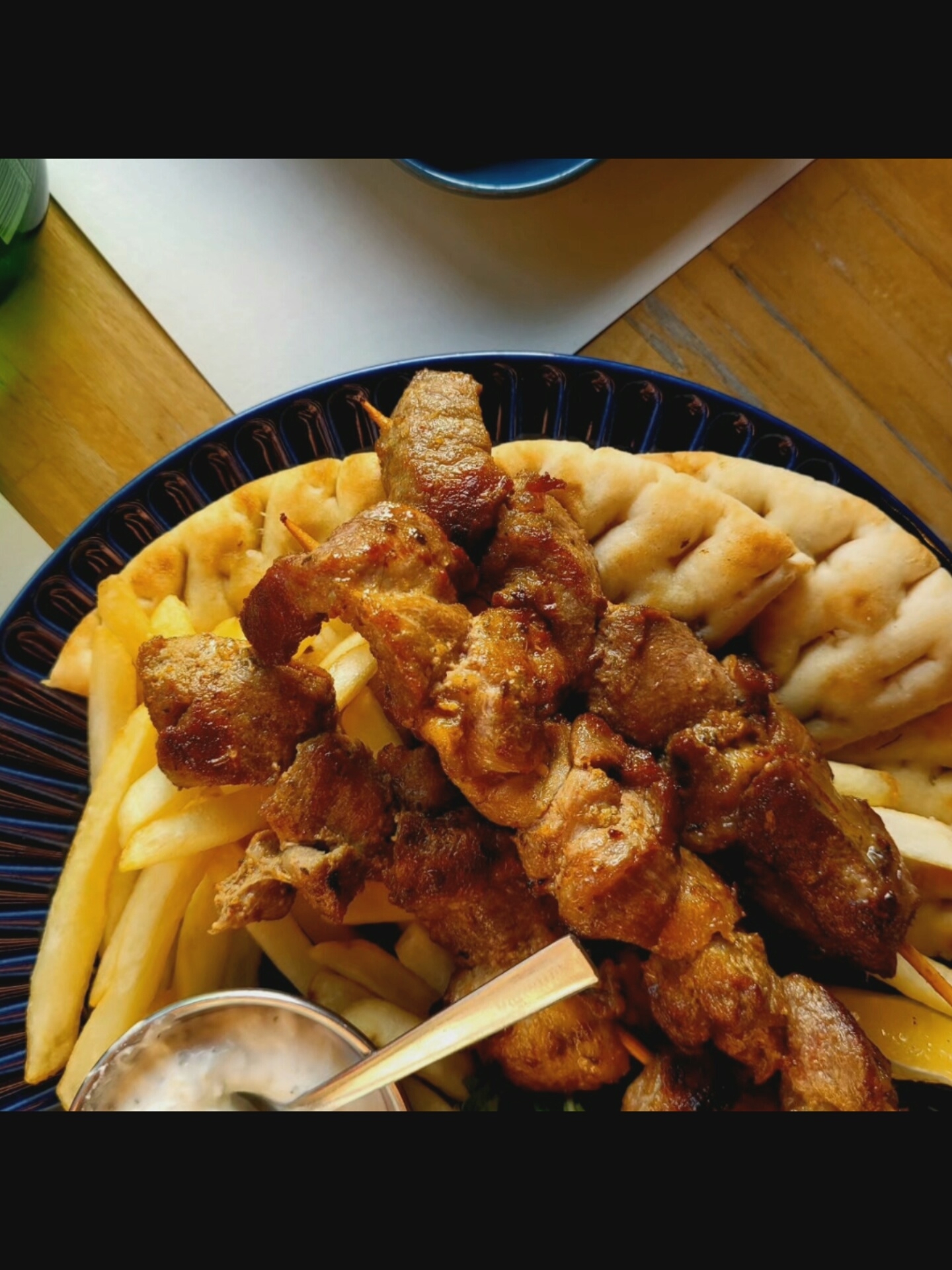 The image shows a close-up of a dish of souvlaki, fries, and pita bread served on a dark blue plate. The souvlaki appears to be made of grilled pork skewers. The fries are golden brown and appear to be thin-cut. The pita bread is flat and slightly puffed. There is a small bowl of white sauce, possibly tzatziki, visible at the bottom left of the plate with a spoon in it. The food is arranged on a wooden table, and a white surface is visible in the background. The lighting suggests it might be daytime. The scene is captured in Bielawska Wieś, Poland.