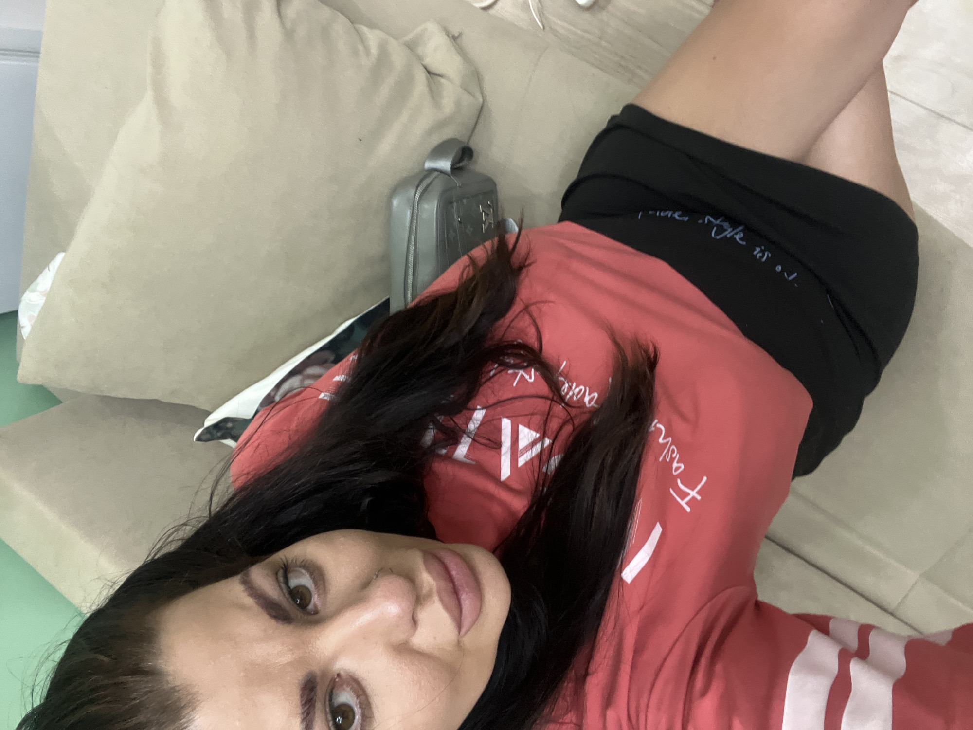 A woman is taking a selfie while sitting on a sofa in a room. She is wearing a red long-sleeved shirt with white stripes on the sleeves and a black skirt. She has dark hair and is looking at the camera. There is a gray purse on the sofa next to her. The room has a light green wall and a beige sofa.