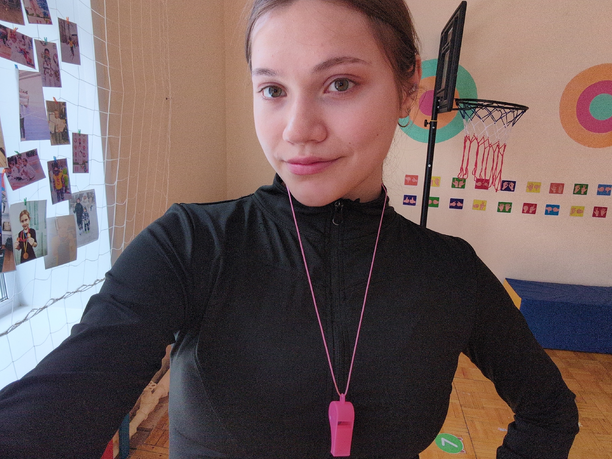 A young woman is captured in a selfie or portrait shot indoors, likely within a gymnasium or activity room in Izhevsk, Russia. She has light brown hair, fair skin, and hazel eyes, looking directly at the camera with a subtle, friendly smile. She wears a black long-sleeved athletic top, and a pink whistle on a pink lanyard is visible around her neck, suggesting a role as a coach, instructor, or referee.
The background displays elements characteristic of a children's sports or activity space. To the left, a white sports net, possibly covering a window, is decorated with numerous small photographs, many featuring children, attached with colorful mini clothes-pins. To the right, a mini basketball hoop with a black backboard and a white net with red loops is set against a wall adorned with two large, concentric circular target designs in orange, pink, and turquoise hues. Below these targets, rows of small, colorful rectangular cards display various hand gestures or symbols. The floor is wooden, resembling a gymnasium, with a green circle containing the number "7" visible. Blue and yellow soft mats or gym equipment are stacked in the lower right background. The bright lighting indicates it is daytime.