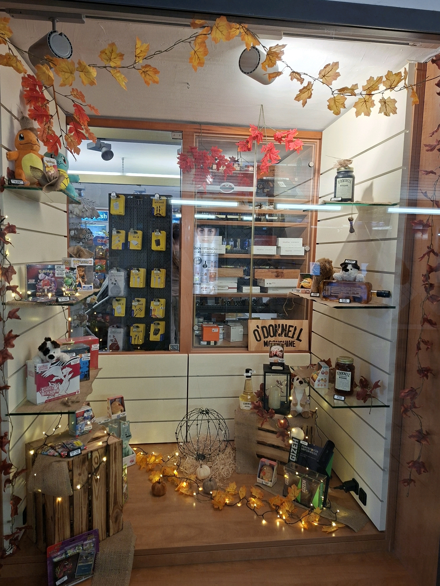 The image shows a shop window display with an autumn theme, featuring a variety of items including plush toys, boxes, bottles, and decorative elements like artificial leaves and string lights. The display is set up on shelves and a wooden crate. In the background, a glass-door cabinet displays more merchandise, possibly cigarettes or related accessories, with some product packaging visible. The shop appears to be located in Koblenz, Germany, as indicated by a sign that reads "O'DONNELL MOONSHINE". The overall atmosphere of the display is festive and seasonal.