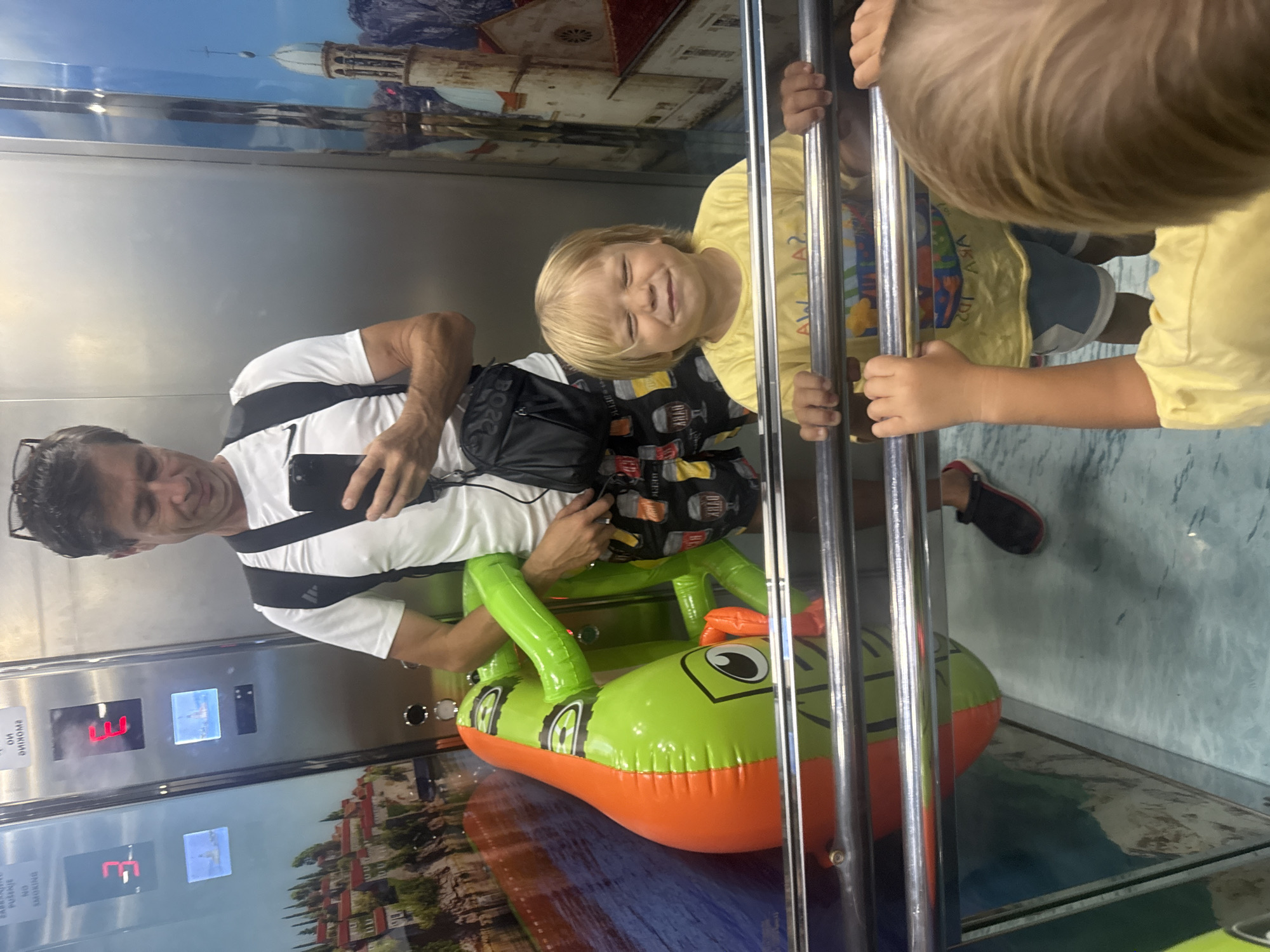 The image shows two children and an adult in an elevator. The adult, a man with dark hair and glasses, is holding a phone and taking a selfie. He is wearing a white t-shirt with a black strap across his chest. A young girl with blonde hair is on the right side of the frame, smiling and holding onto the elevator railing. She is wearing a yellow shirt. Behind her, another child is partially visible, also in a yellow shirt. The elevator is decorated with murals on the walls, depicting scenic European-style buildings and landscapes. On the floor of the elevator is a large, inflatable green and orange crocodile. The elevator buttons are visible on the left, with the number "3" displayed. The context of Boreti, Montenegro, suggests the scene is likely in a tourist area. The overall mood seems cheerful and playful, with the children appearing happy.