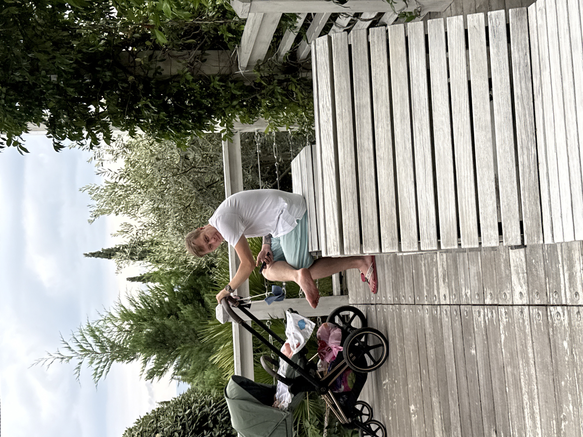 A man is sitting on a wooden bench on a wooden deck next to a baby stroller. He is wearing a white t-shirt and light blue shorts, and flip-flops. He is holding a mobile phone in his right hand and looking at the camera. The baby stroller is green and is empty except for a blanket and some pink plastic bags. The deck is made of weathered wooden planks and is surrounded by lush greenery. The sky is cloudy. The scene appears to be in an outdoor recreational area, possibly a park or a resort. The location is listed as Yalta, Ukraine.