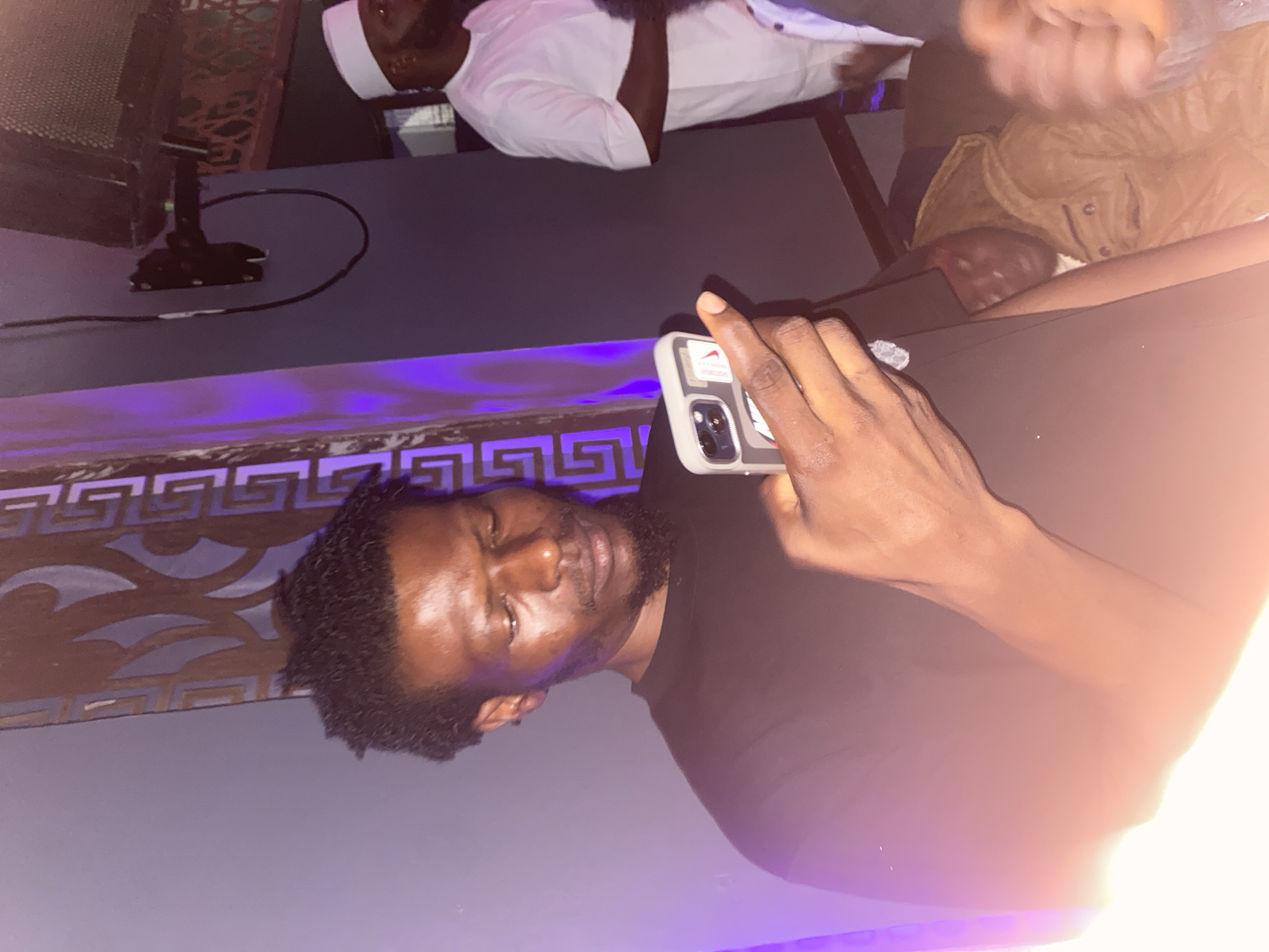 A man in a black t-shirt holds a smartphone in his right hand and looks at the screen, his head tilted down. He has dark, curly hair and a goatee. The background features purple lighting and a decorative wall panel. To the right, another man wearing a white traditional outfit and a cap is visible. The scene appears to be indoors, possibly in a club or event space, with a speaker mounted on the wall above. The overall atmosphere is dim, illuminated by ambient purple and white light.