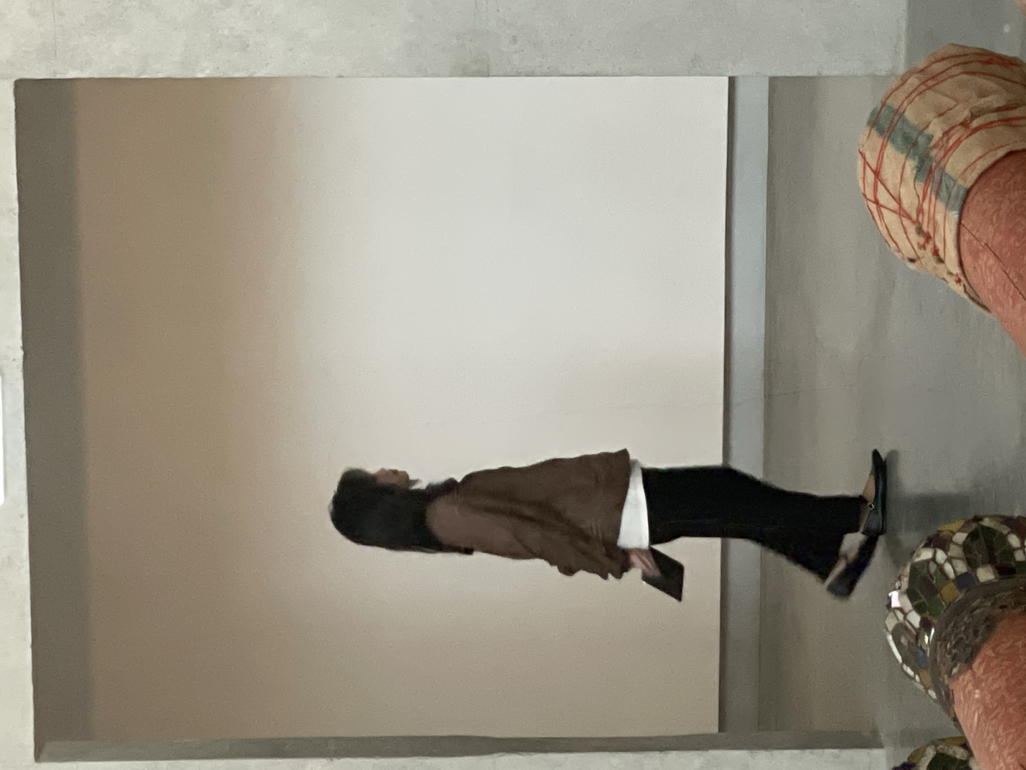 A woman is seen standing in a room with plain white walls. She is wearing a brown jacket, a white shirt, and black pants. She is holding a tablet or electronic device. In the foreground, partially visible, are some decorative items, one with a patchwork pattern and another with a mosaic design. The location is unknown and cannot be determined as being in Bangkok, Thailand.