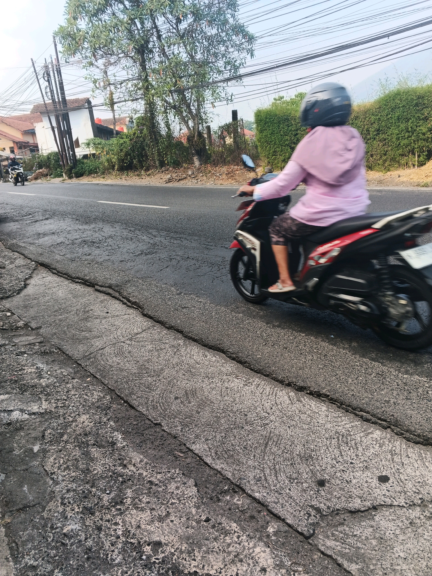 A person wearing a pink hooded shirt and a black helmet rides a red and black motorcycle on a paved road in Ungaran, Indonesia. The motorcycle is in motion, indicated by motion blur. The road has a cracked and worn asphalt surface, with a concrete curb on the left side. In the background, there are houses, trees, and power lines. The sky is overcast. The scene appears to be during daylight hours.