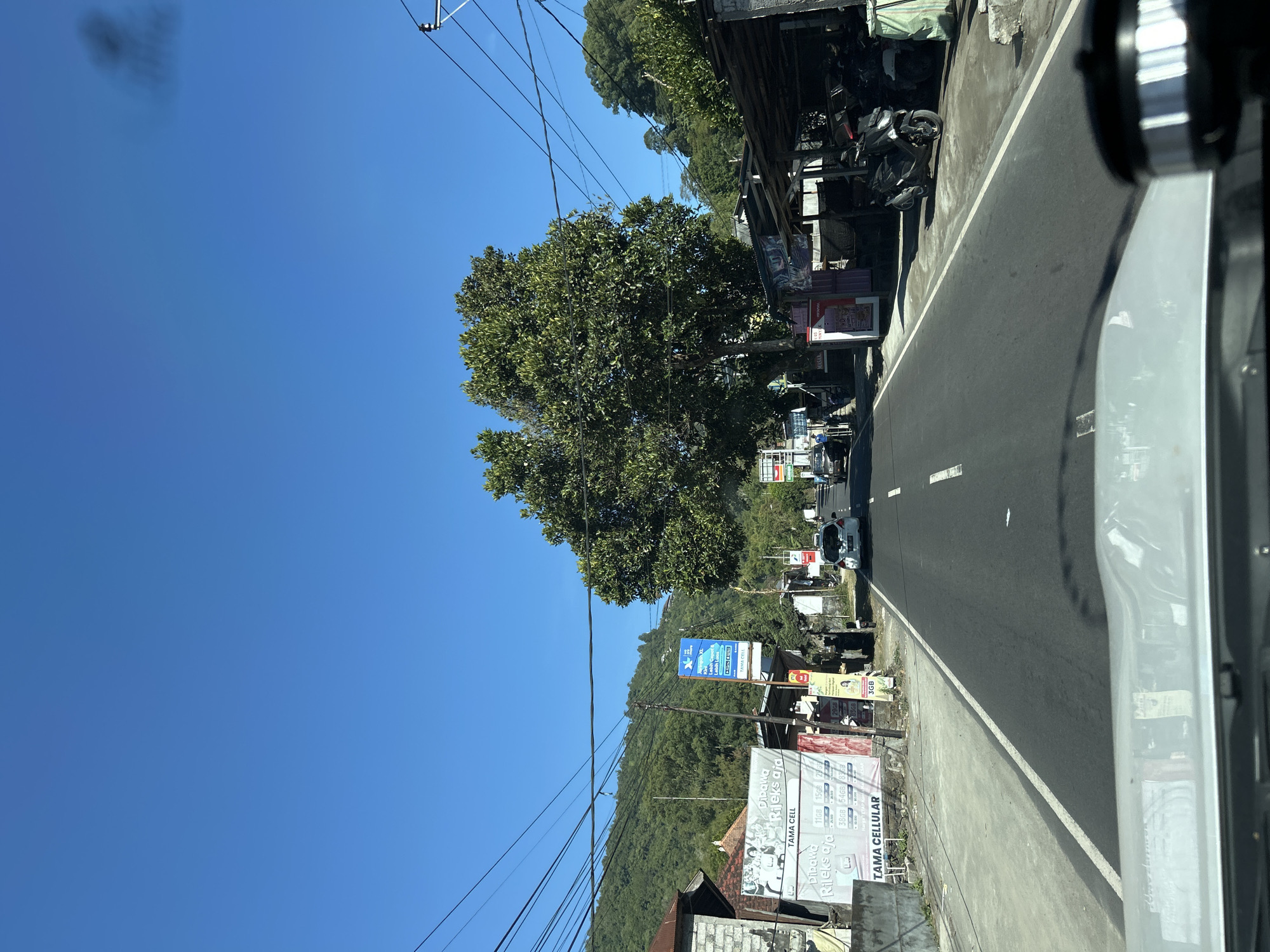 The image shows a straight, paved road in Kintamani, Indonesia, under a clear blue sky. The road has a white line marking the center. On the left side of the road, there is a low, white building with a brick wall, and a large banner advertising "TAMA CELLULAR" with various data package prices. Further down the road on the left, a tall, blue advertisement for XL Axiata is visible. There are several parked motorcycles and scooters on the right side of the road, under a simple wooden shelter with a corrugated metal roof. Behind the shelter, more parked motorcycles are visible. Lush green trees and vegetation cover the rolling hills in the background. The scene appears to be a street in a rural or semi-rural area, possibly a small town, with commercial activity related to a cellular provider. A white car is visible driving away from the camera on the right side of the road, and another vehicle is further ahead. The foreground shows the dashboard and windshield of a vehicle from which the photo was taken.