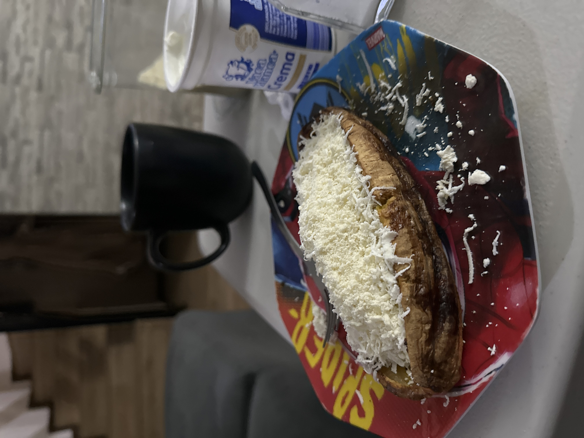 An indoor, close-up shot displays a cooked plantain, likely fried or baked, generously covered with finely grated white cheese. The plantain sits on a colorful, hexagonal-shaped plate adorned with Marvel characters, where "SPIDER" and "MARVEL" text is visible. A black fork is partially placed on the plate, near the food. In the blurred background, a white plastic container of "Crema" is present, with the word "Crema" clearly legible on its label, alongside other unreadable text. A black mug is also seen behind the food. The deeper background appears to be an interior space, with indistinct wooden elements, possibly stairs or shelving, and a grey piece of furniture. The image captures a casual meal or snack, possibly a common dish found in Puebla, Mexico.