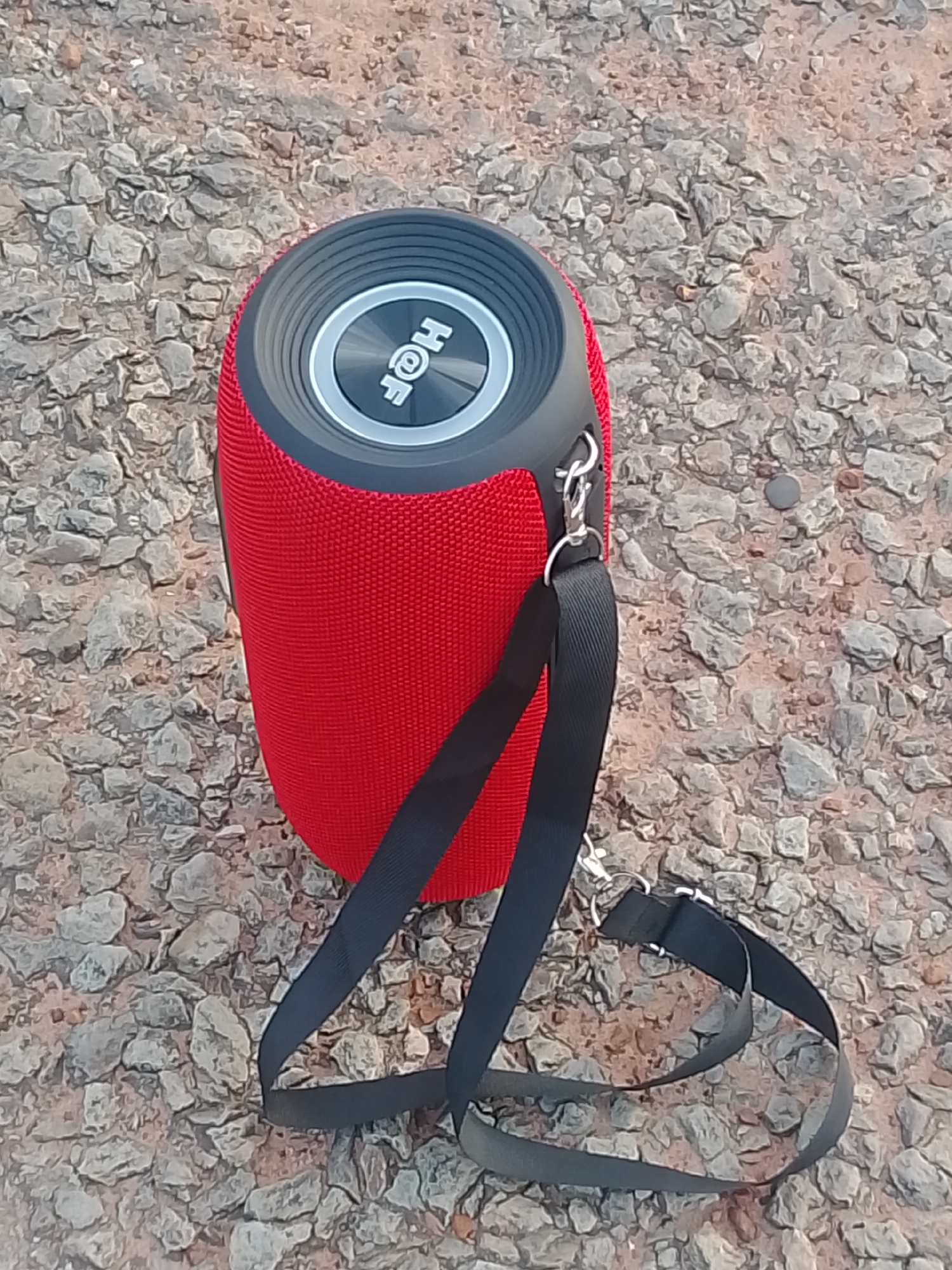 An outdoor, eye-level, close-up shot focuses on a red and black portable Bluetooth speaker lying on a bed of small, grey stones and dirt. The speaker is cylindrical with a textured red fabric body and a black top with a silver ring encircling a circular logo that reads "HQF". A black strap is attached to the speaker by metal clips. The strap is partially unwound, with one end leading away from the speaker and the other end looping around the speaker and resting on the ground. The background is an unfocused expanse of stones and dirt, suggesting an outdoor, possibly unpaved, location. There are no people or animals visible in the image.