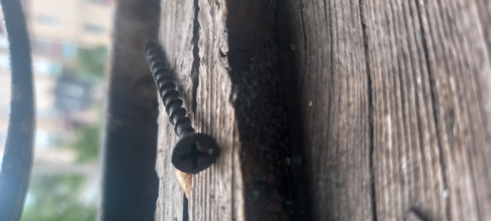 A close-up shot depicts a black screw partially embedded in weathered wooden boards. The wood grain is visible, and there is evidence of cracks and aging. In the blurry background, an urban landscape hints at a building or apartment complex, suggesting an outdoor setting in a city. The picture was taken in Kryvyi Rih, Ukraine. There are no people or interactions visible in the image.