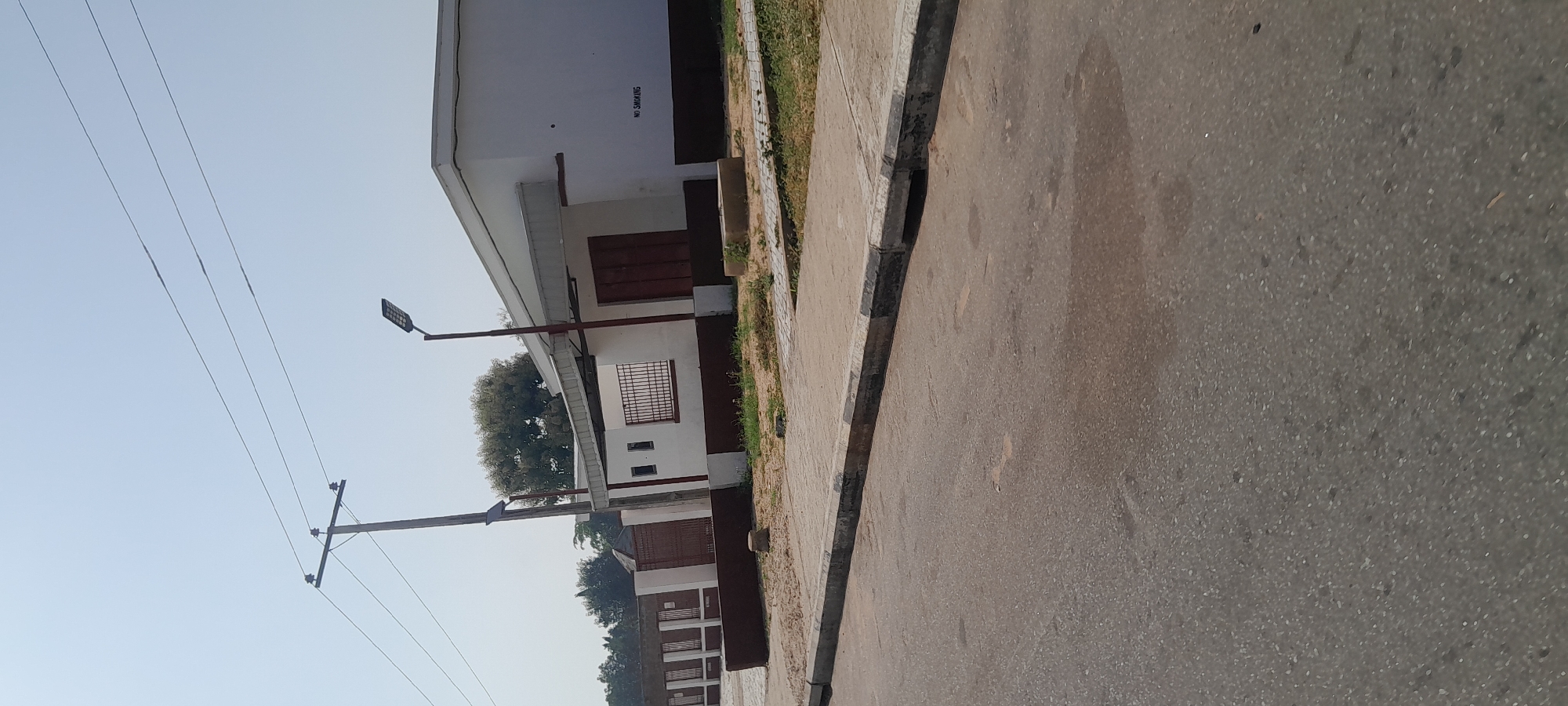 The image depicts a street scene in Jalingo, Nigeria, under a clear sky. The foreground is dominated by an asphalt road with a concrete curb and sidewalk. Beyond the sidewalk is a building with a white facade and a brown base. The building has multiple windows with metal bars and large brown double doors. A street lamp with an LED fixture is mounted on a pole in front of the building. To the left of the building, a utility pole with electrical wires is visible. Further to the left, a fence made of brown metal bars and white posts separates the property from the street. Trees are visible behind the fence and the building. A sign with "NO SMOKING" written in black is affixed to the right side of the building. The environment appears to be an urban or suburban area. No people are visible in the image.