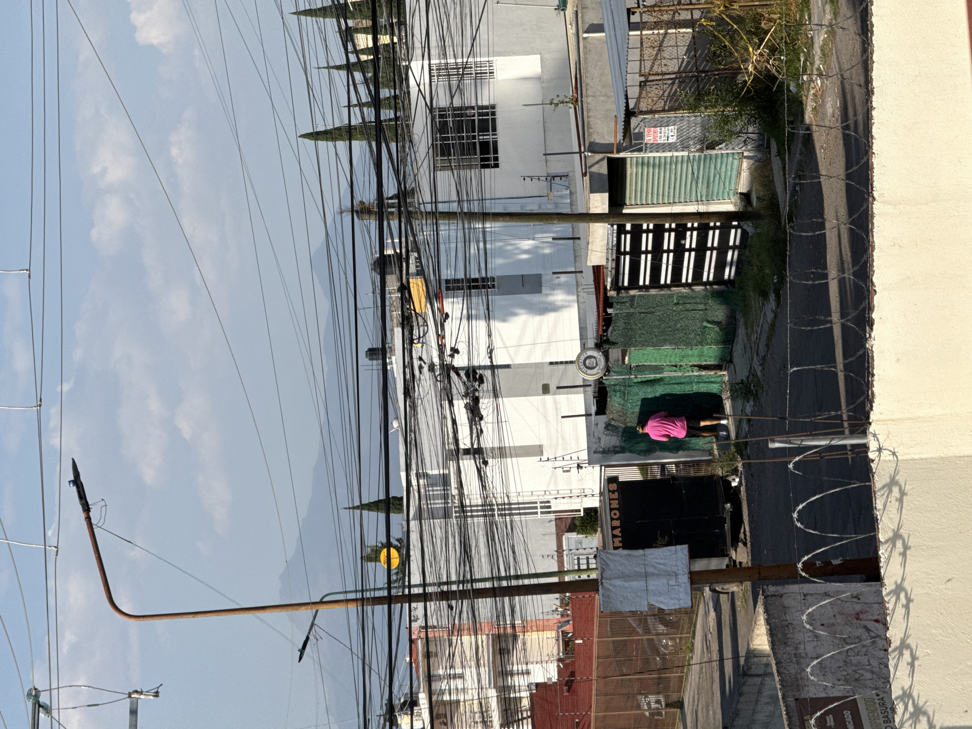 The image shows a street scene in Puebla, Mexico. Over tangled power lines is a view of buildings, including some white and red-brick structures, with tall thin trees standing near the buildings. On the right side, along the sidewalk, a person wearing a pink shirt is lying on a green mat. A fence with barbed wire runs along the side of the street. Signs and a lamppost are also visible along the sidewalk.