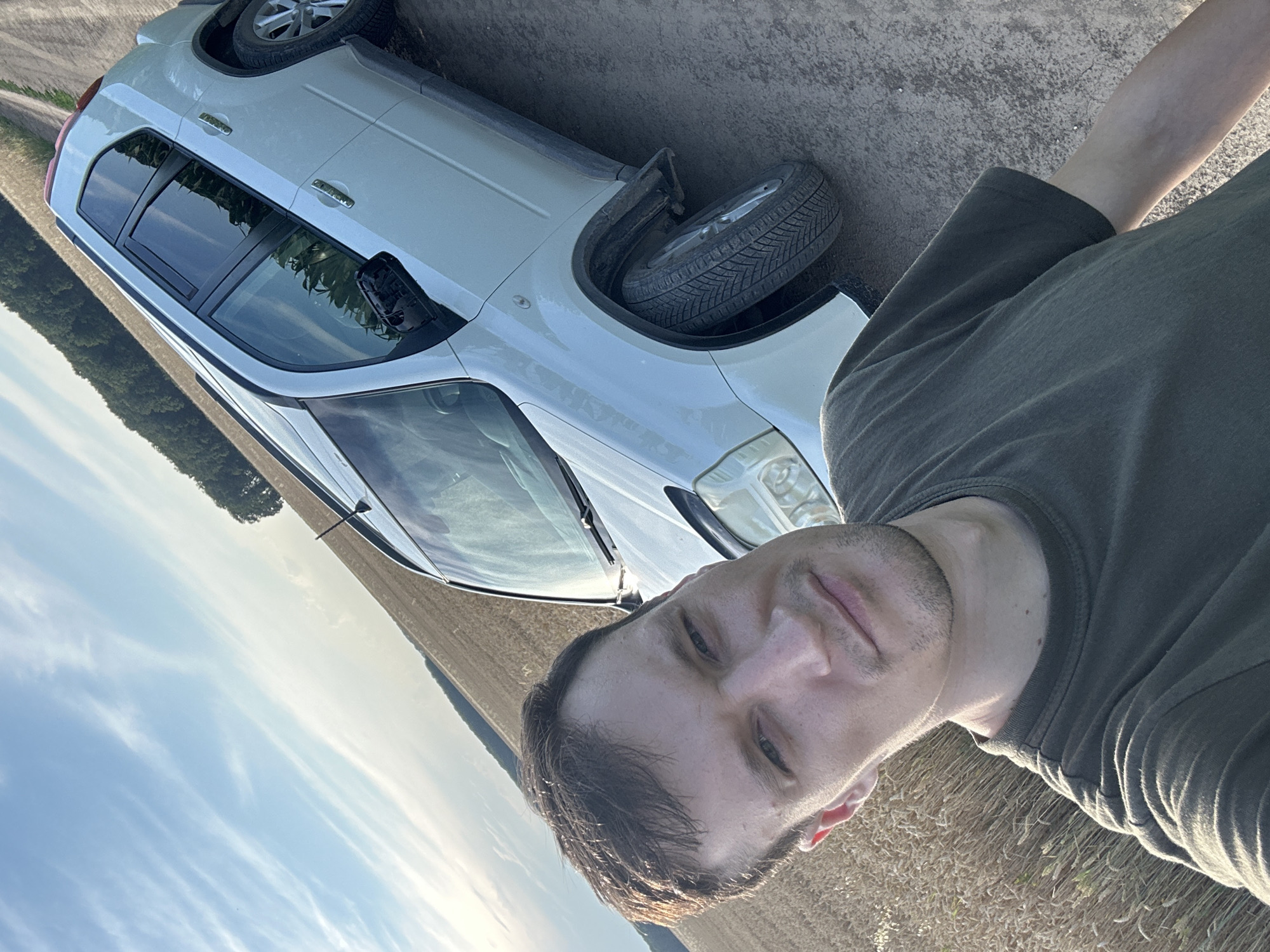 A man in a green t-shirt is taking a selfie next to a white Nissan X-Trail SUV parked on a dirt road. The car is positioned at an angle to the right, with its front left wheel slightly turned. The man's face is partially visible in the foreground, looking directly at the camera. Behind the car, a field of ripe wheat stretches towards a treeline, and beyond that, a clear blue sky with wispy clouds. The image appears to have been taken during daylight, likely in the late afternoon due to the quality of light. The location is the village of Slobidka-Zalisetska, Lviv Oblast, Ukraine.