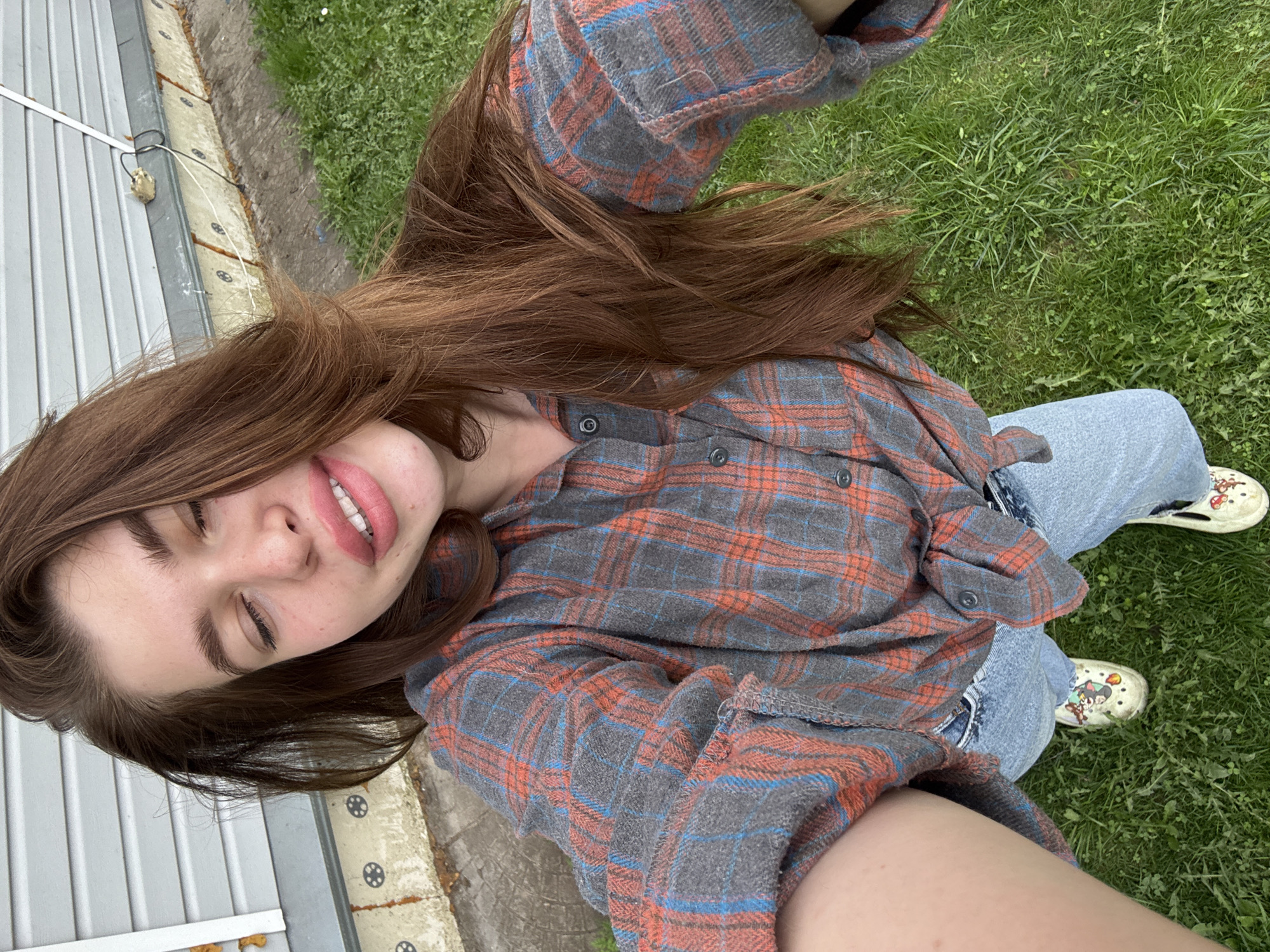 A young woman is lying on a patch of grass next to a building. She is wearing a plaid shirt and jeans. She is wearing crocs on her feet. She is smiling with her eyes closed. The location is Мещёры, Russia.