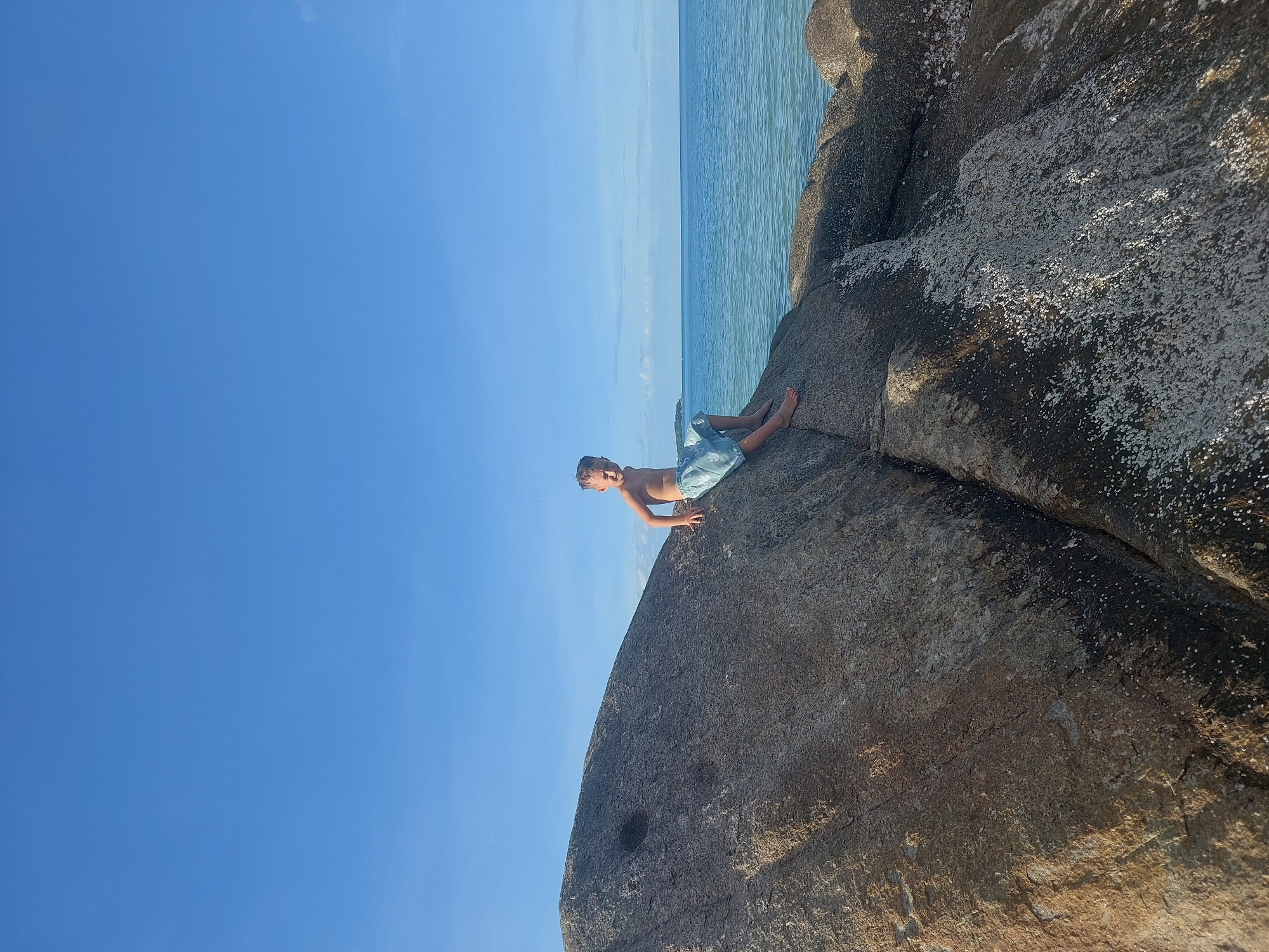 A young boy is lying on a large, grey, textured rock at the edge of the water. He is wearing light blue patterned swim shorts and is shirtless. His body is angled downwards, with his head propped up on his hands, looking towards the camera. His expression appears relaxed and happy. Behind him, the clear blue ocean stretches out to meet a pale blue sky with wispy white clouds. The sun is shining brightly, casting shadows on the rock and illuminating the water. The rock surface is rough and uneven, with small white flecks scattered across it, possibly barnacles or shell fragments. The scene suggests a warm, sunny day at the beach in Baan Chaweng Noi, Thailand, with the boy enjoying a moment of leisure by the sea. The overall mood is peaceful and idyllic. There is no visible text in the image.