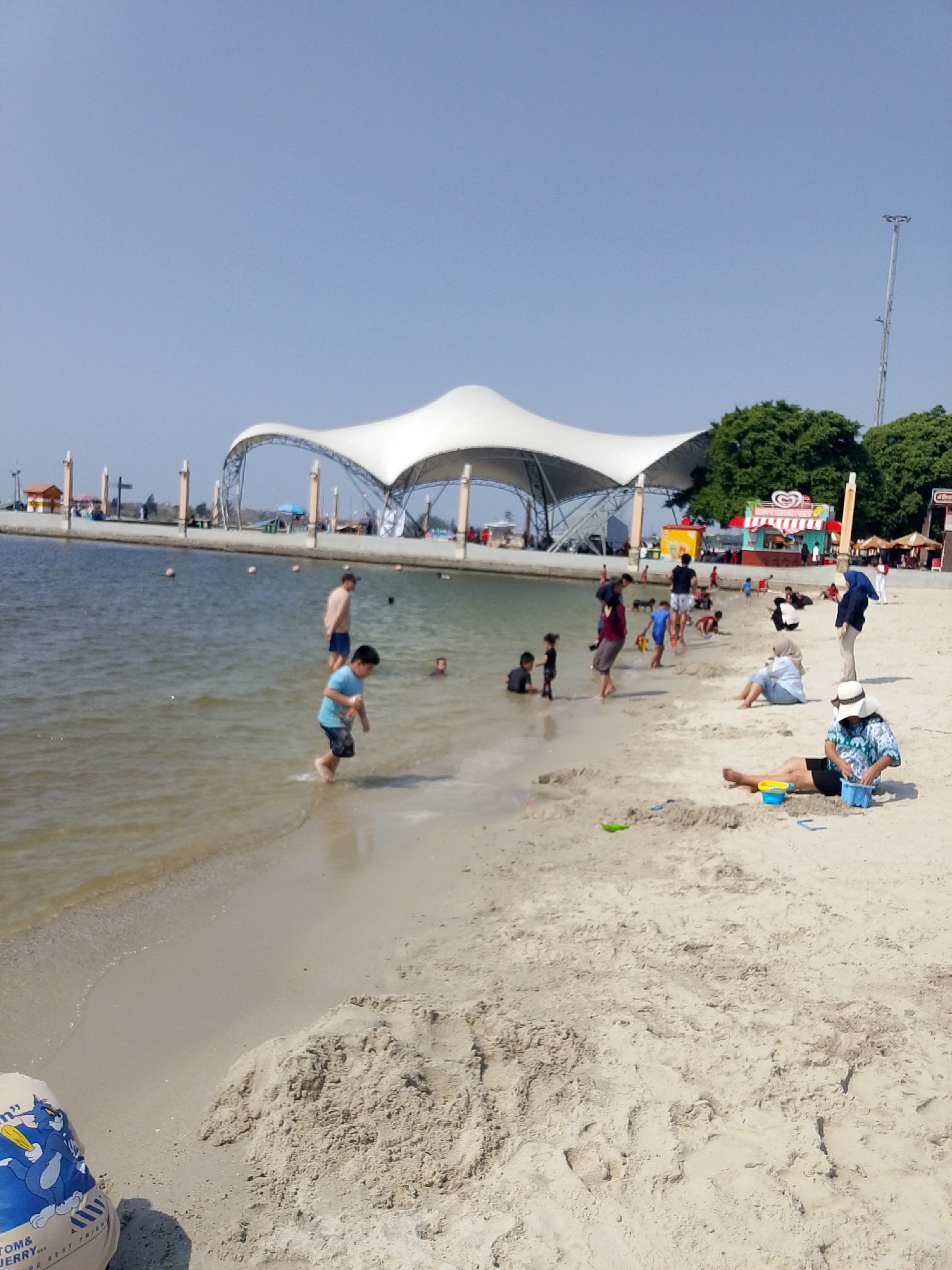 This is a wide-angle, eye-level shot of Ancol Beach, North Jakarta, Indonesia on a clear, sunny day. The foreground is dominated by a sandy beach with some footprints and a pile of sand, possibly from children playing. A blue sand bucket and a yellow shovel are visible in the sand near a seated person. Further into the image, there is a calm, shallow body of water with several people wading and playing. A young boy in a blue shirt and dark shorts is in the shallow water, walking towards the beach. A man in a light-colored shirt and dark shorts stands further back in the water. In the middle ground, the beach continues with more people, including a woman in a light blue top and a headscarf sitting on the sand, and another person in a white hat and patterned top also sitting on the sand and playing with a blue bucket. In the background, there is a large, white, modern-looking canopy structure, likely part of a recreational facility. Beyond that, there are some trees, food stalls, and other beachgoers. The overall scene depicts a relaxed atmosphere with people enjoying a day at the beach.