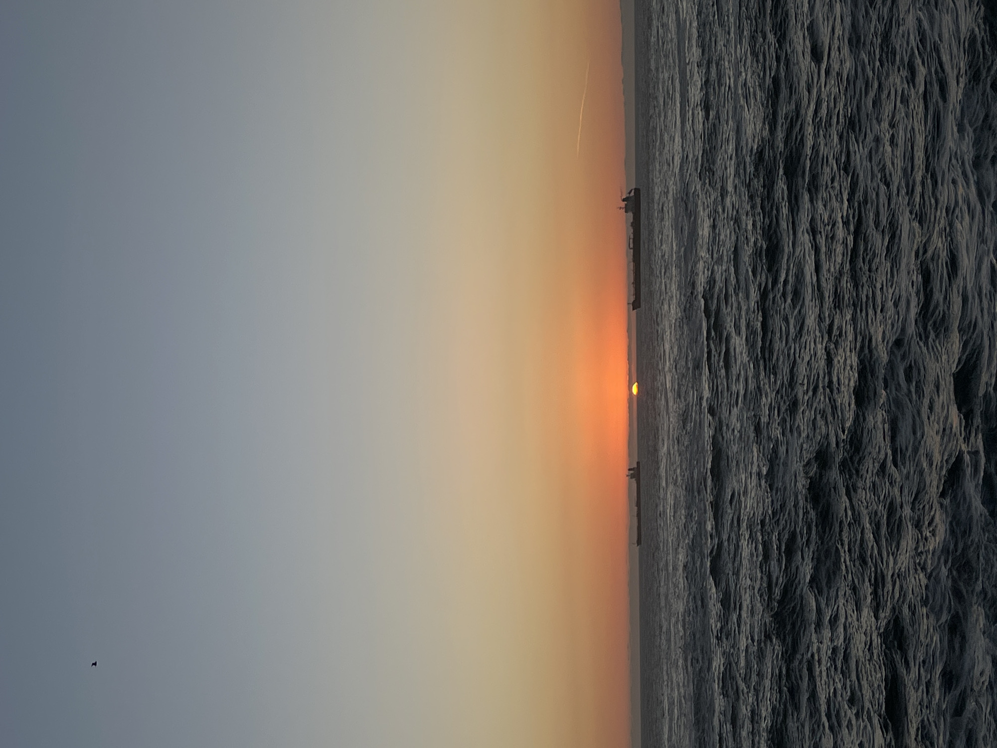 The image depicts a sunset over the ocean, likely in Arica, Chile. The sun is positioned on the horizon, partially obscured by vessels, creating a gradient of orange and yellow hues in the sky. To the left of the sun, the sky transitions to a light blue. The ocean's surface, to the right of the horizon, appears textured and dark, reflecting the fading light. There is a contrail stretching out on the top right. A bird is flying on the bottom left.