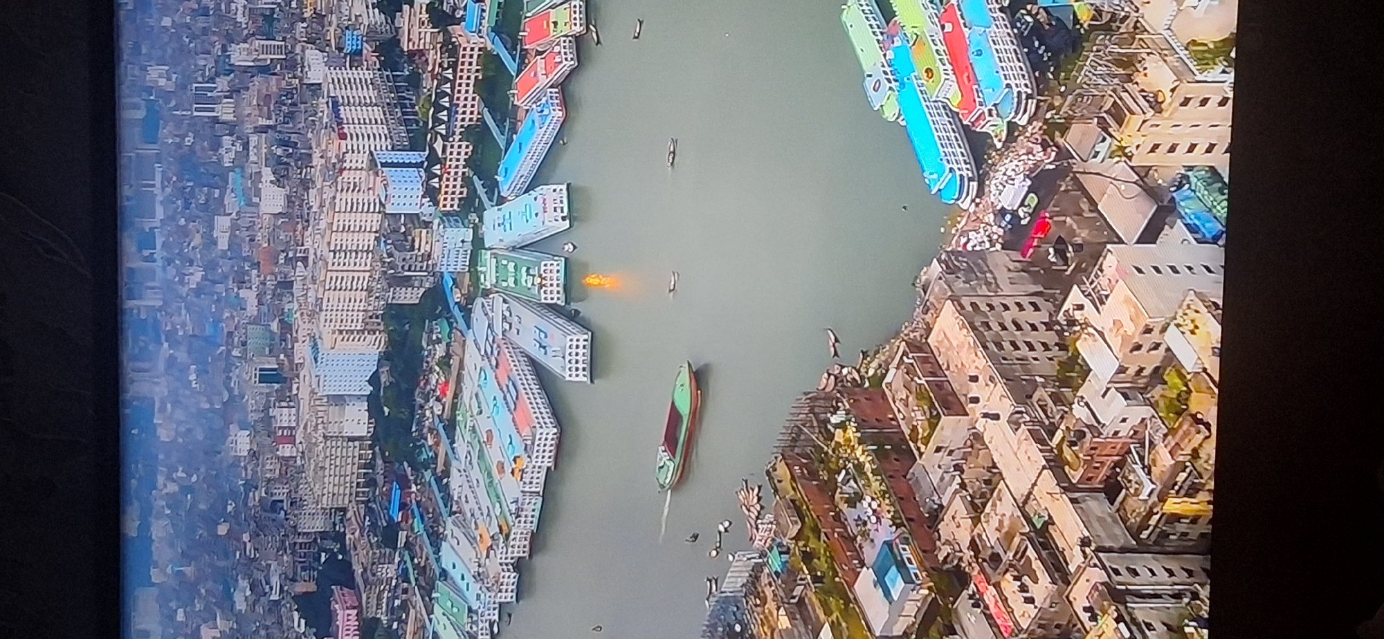 The image is an aerial view of a densely populated urban area with a prominent river or waterway. Along the river are several long, multi-decked passenger vessels, some colored green, blue, and red. In the foreground, closer to the camera, is a cityscape composed of numerous buildings of varying heights and architectural styles, many appearing to be apartment blocks or commercial structures. Some buildings show signs of wear or unfinished construction. A cargo barge is moving in the waterway. The overall impression is of a bustling port city with extensive residential and industrial areas. This scene is not located in Bila Tserkva, Ukraine.