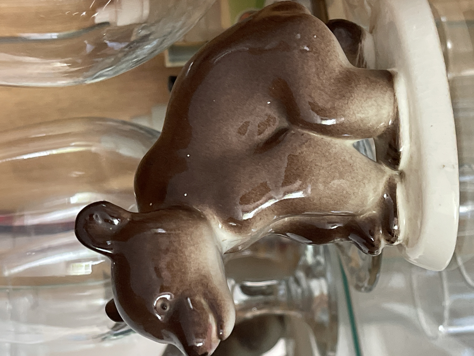 The image features a close-up of a small, brown, ceramic bear figurine. The figurine is shiny and appears to be glazed. It is positioned on top of a white, round base, and the bear is depicted in a crouching or sitting position, with its head facing downwards and to the left. Its ears are visible, as is one eye, which is painted as a small black dot with a white highlight. The figurine is glossy, reflecting the light and the surrounding environment.
The background is blurred and out of focus, but appears to be composed of glass objects, possibly jars or containers, with reflections and refractions of light. The overall impression is that the figurine is placed among other items, perhaps on a shelf or in a display. The lighting seems to be artificial, and the colors are warm and muted. There is no visible text in the image. The scene does not provide cues about the time of day, weather, or a specific location beyond what can be inferred from the objects present.