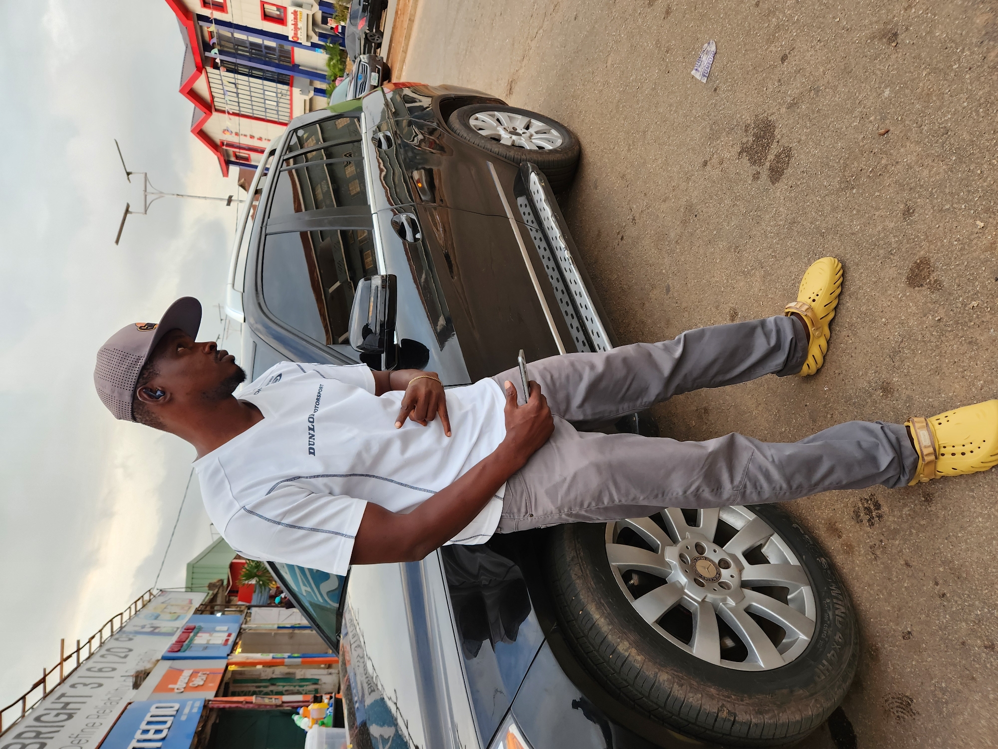 A man stands next to a black Mercedes-Benz SUV on a paved street. He is wearing a white t-shirt with a blue logo, grey pants, a grey baseball cap, and yellow clogs. He is holding a smartphone in his right hand and has his left hand raised in a gesture. The SUV has silver rims and running boards. In the background, there are buildings with red and blue accents, streetlights, and other vehicles. The sky is overcast.