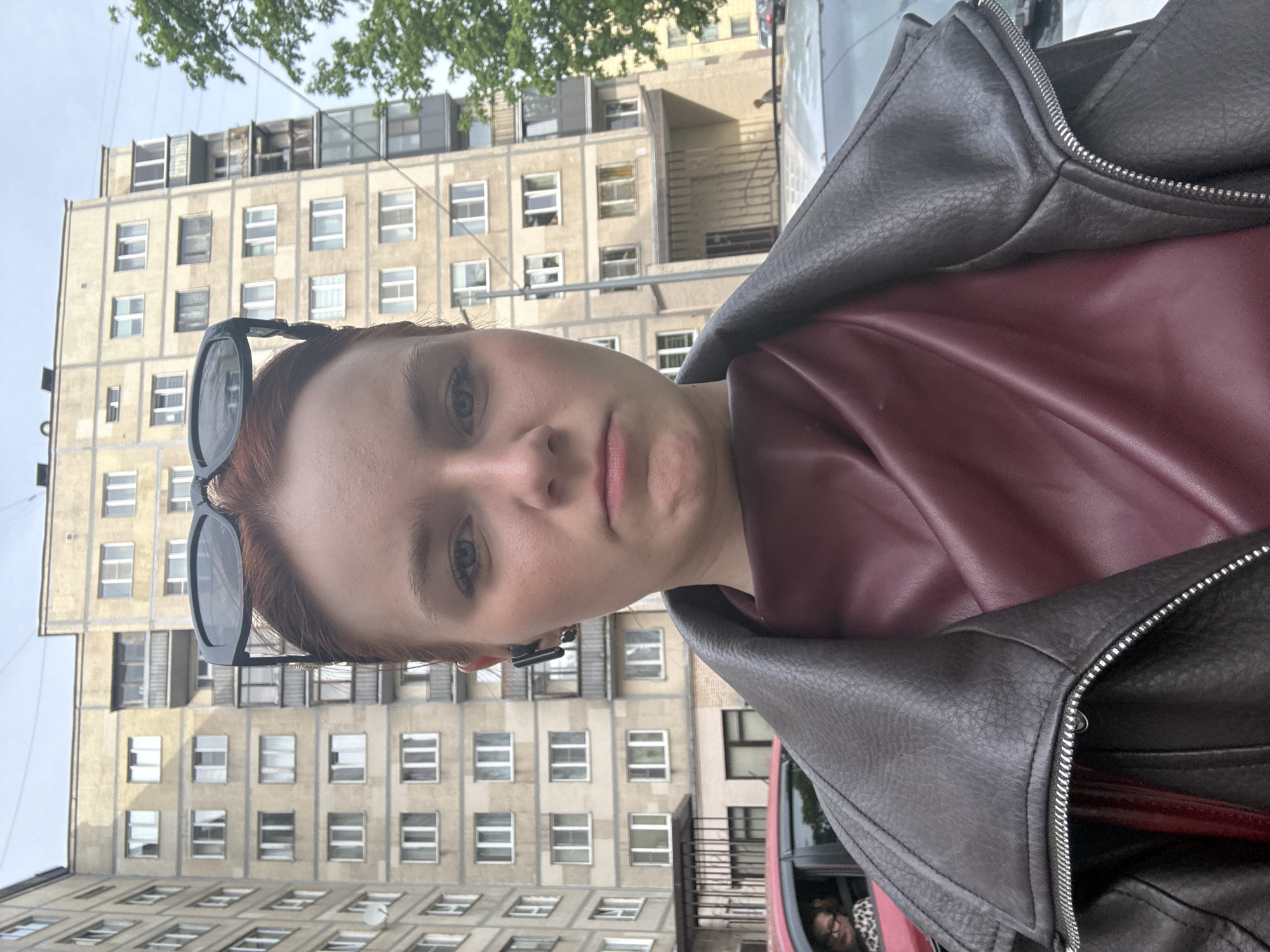 A woman poses close to the camera with her head tilted, and a serious expression on her face. She is wearing black sunglasses on her head, a dark brown leather jacket, and a burgundy-colored shirt. Behind her is a tall, Soviet-era residential building with multiple rows of windows. In the reflection of a car window in the lower right corner of the image, another person can be seen. Based on the architectural style of the building and the overall environment, the location is likely in Saint Petersburg, Russia.