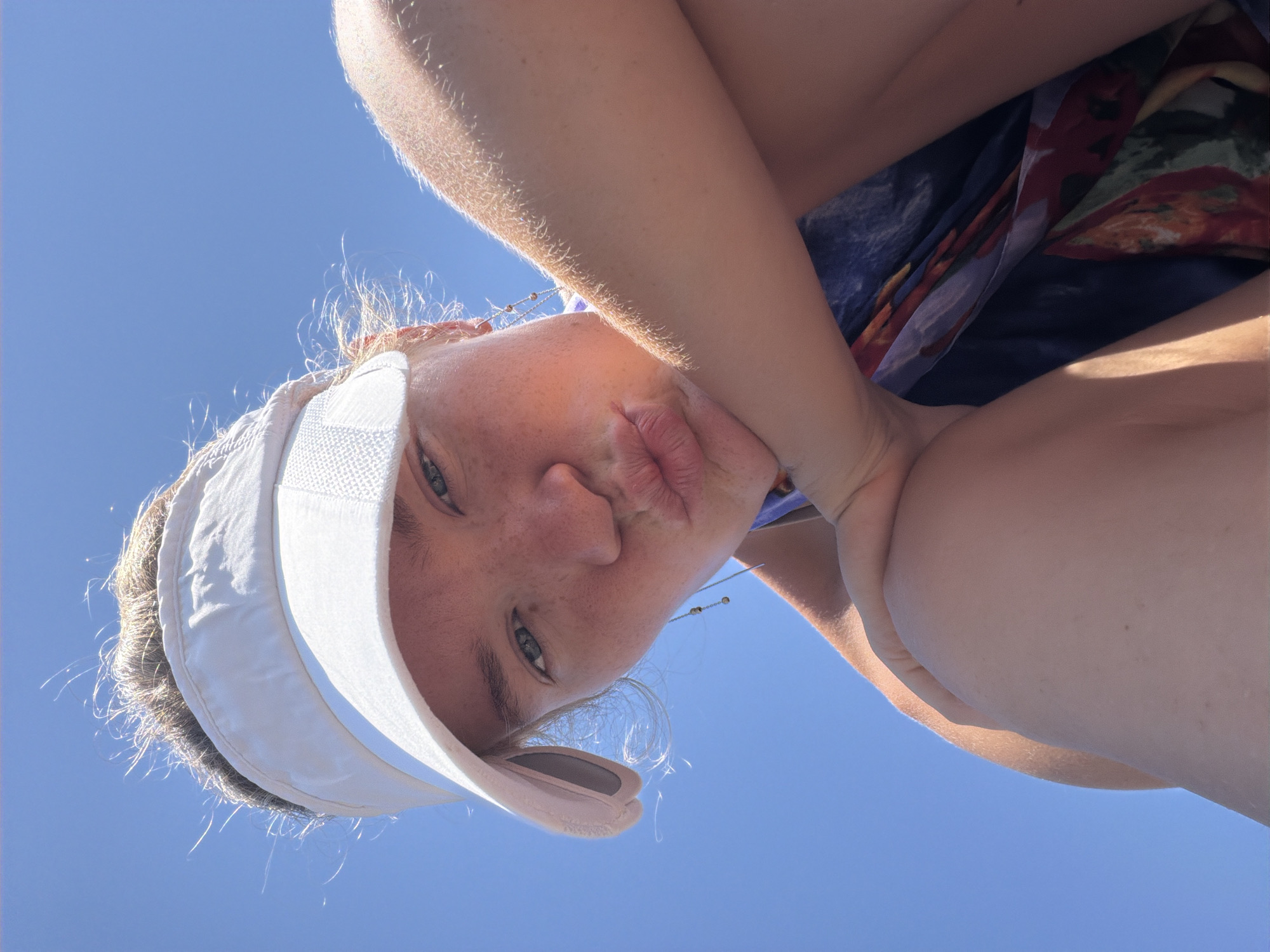A young person is captured in a low-angle, upside-down selfie against a clear blue sky. They are wearing a white visor, which casts a slight shadow on their forehead. Their eyes are open and looking towards the camera, and their lips are pursed in a playful pout. Freckles are visible on their nose and cheeks. Fine, light-colored hairs are catching the sunlight around their head. One arm is raised, with the hand resting near their chin, and the other arm is bent at the elbow. A colorful, patterned fabric, possibly part of clothing or a scarf, is visible beneath their arm. The overall impression is one of a casual, sunny moment, possibly at the beach or outdoors, with the bright sky suggesting daytime and fair weather. There is no visible text in the image.