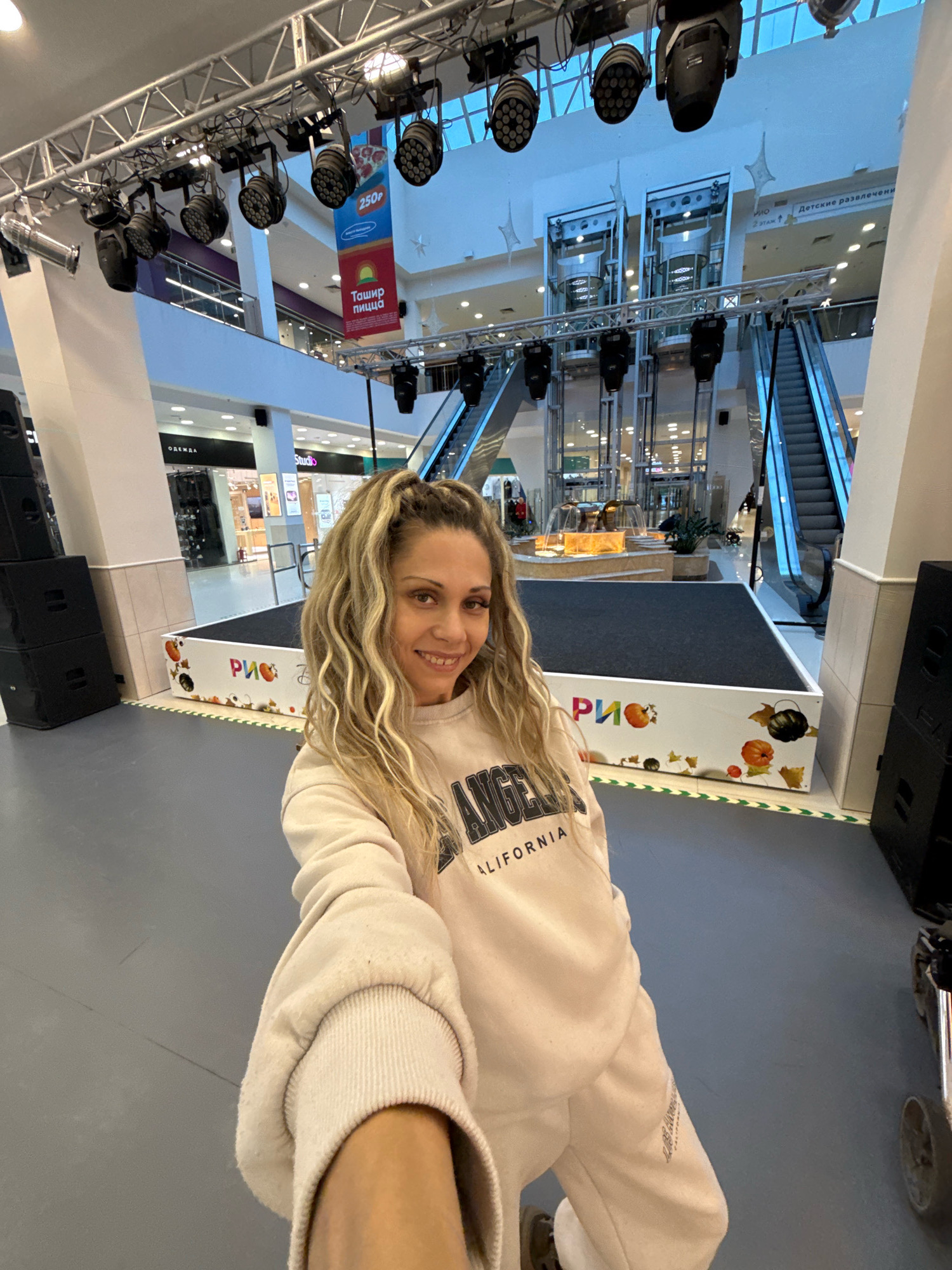 A woman with blonde curly hair pulled into a ponytail takes a selfie in a shopping mall. She is wearing a light-colored sweatshirt and sweatpants. She is standing on a grey floor, with a stage decorated with pumpkins and the word "РИО" in front of her. In the background, there are escalators, elevators, shops, and lighting equipment suspended from the ceiling. The scene is well-lit, and the overall atmosphere appears to be indoors within a commercial or entertainment venue. The location is identified as Rusanovo, Russia.