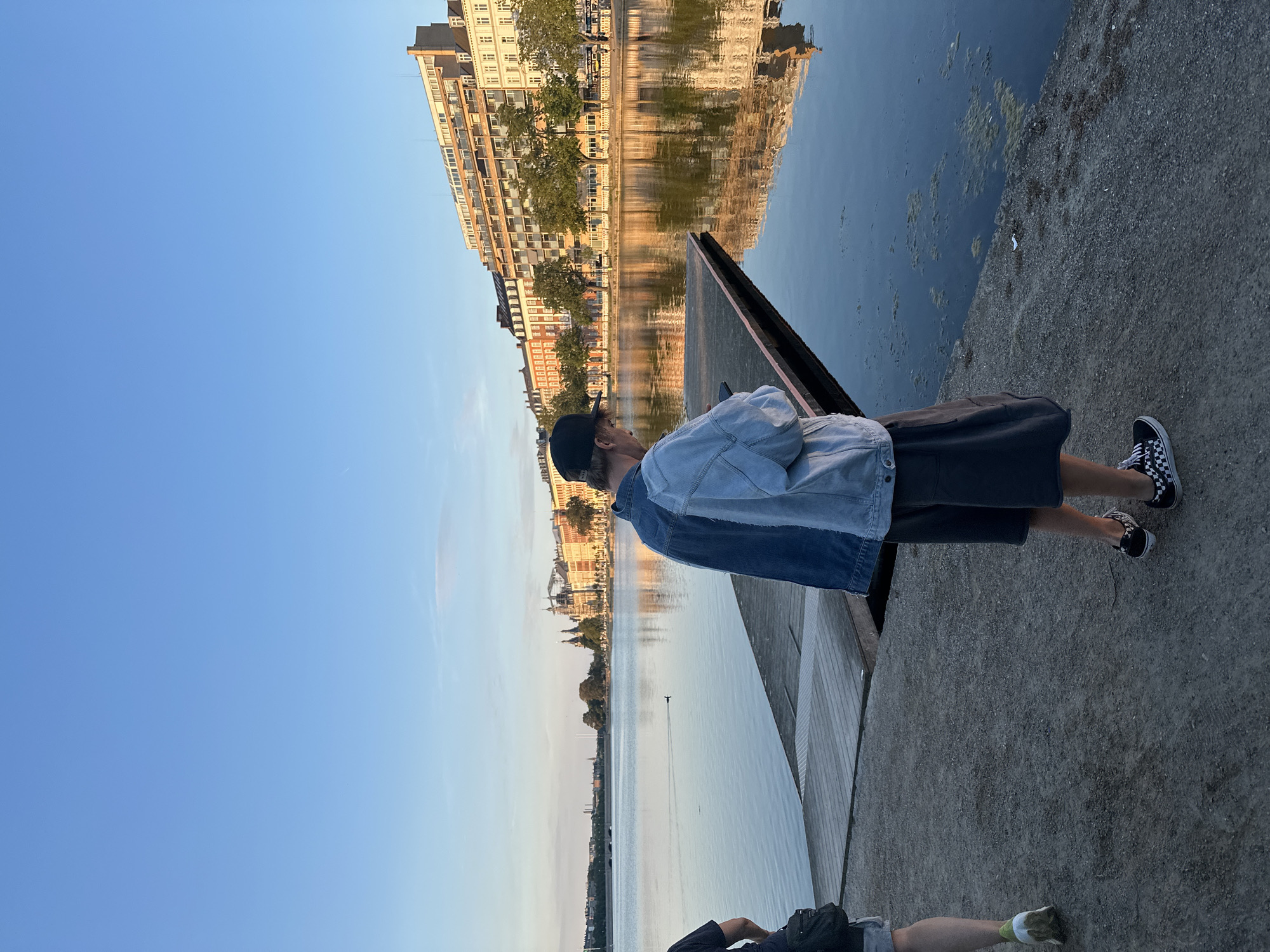 The image depicts a waterfront scene in Copenhagen, Denmark, likely in the vicinity of a canal or harbor. In the foreground, two individuals stand on a paved area next to the water. One person, facing away from the camera, is wearing a denim jacket, dark shorts, and checkered sneakers, and is looking at their phone. Another person is partially visible on the left, walking away from the camera.
The water's surface is calm, reflecting the clear blue sky and the buildings lining the opposite bank. These buildings are multi-story, with a mix of architectural styles, and appear to be residential or commercial properties. Trees line the waterfront, and the golden hues of the late afternoon or early evening sun cast a warm glow on the scene, illuminating the reflections in the water. A small duck is visible swimming in the canal. A wooden pier extends into the water. The overall atmosphere is serene, with a sense of urban tranquility.