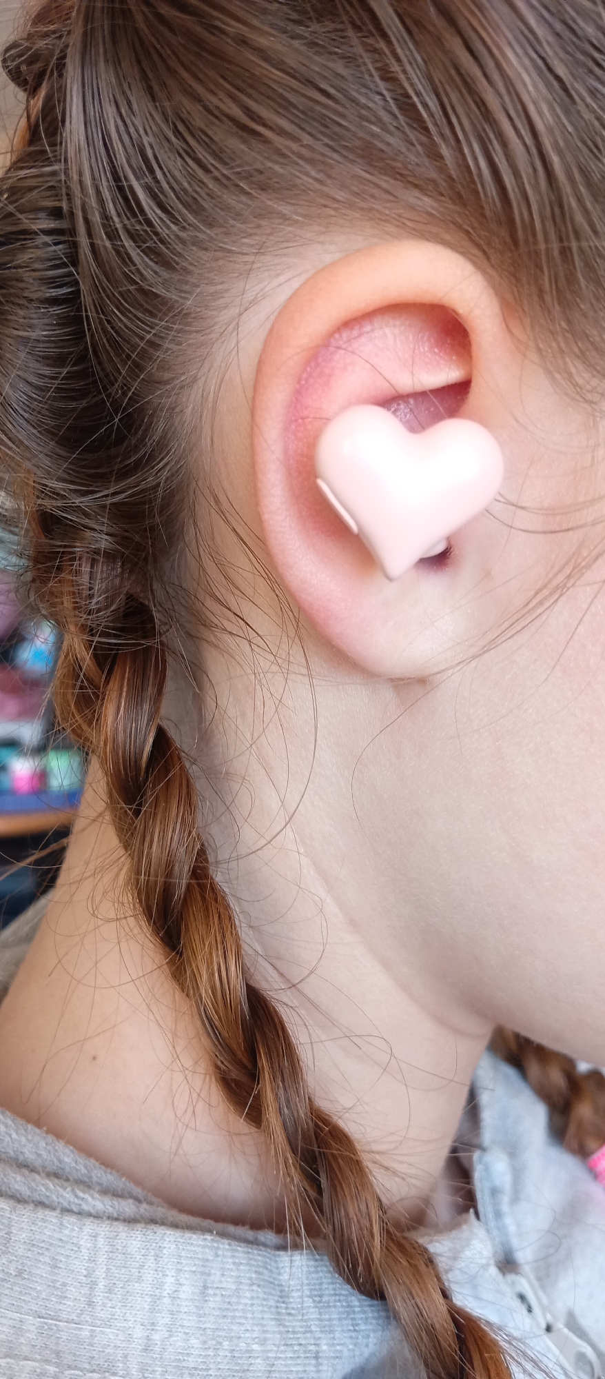 A close-up shot depicts a person's ear with a pink heart-shaped object, likely an earbud, inserted into the ear canal. The person has brown hair styled in braids. The background is blurred, showing hints of other objects and colors. The location is Unknown, Unknown.