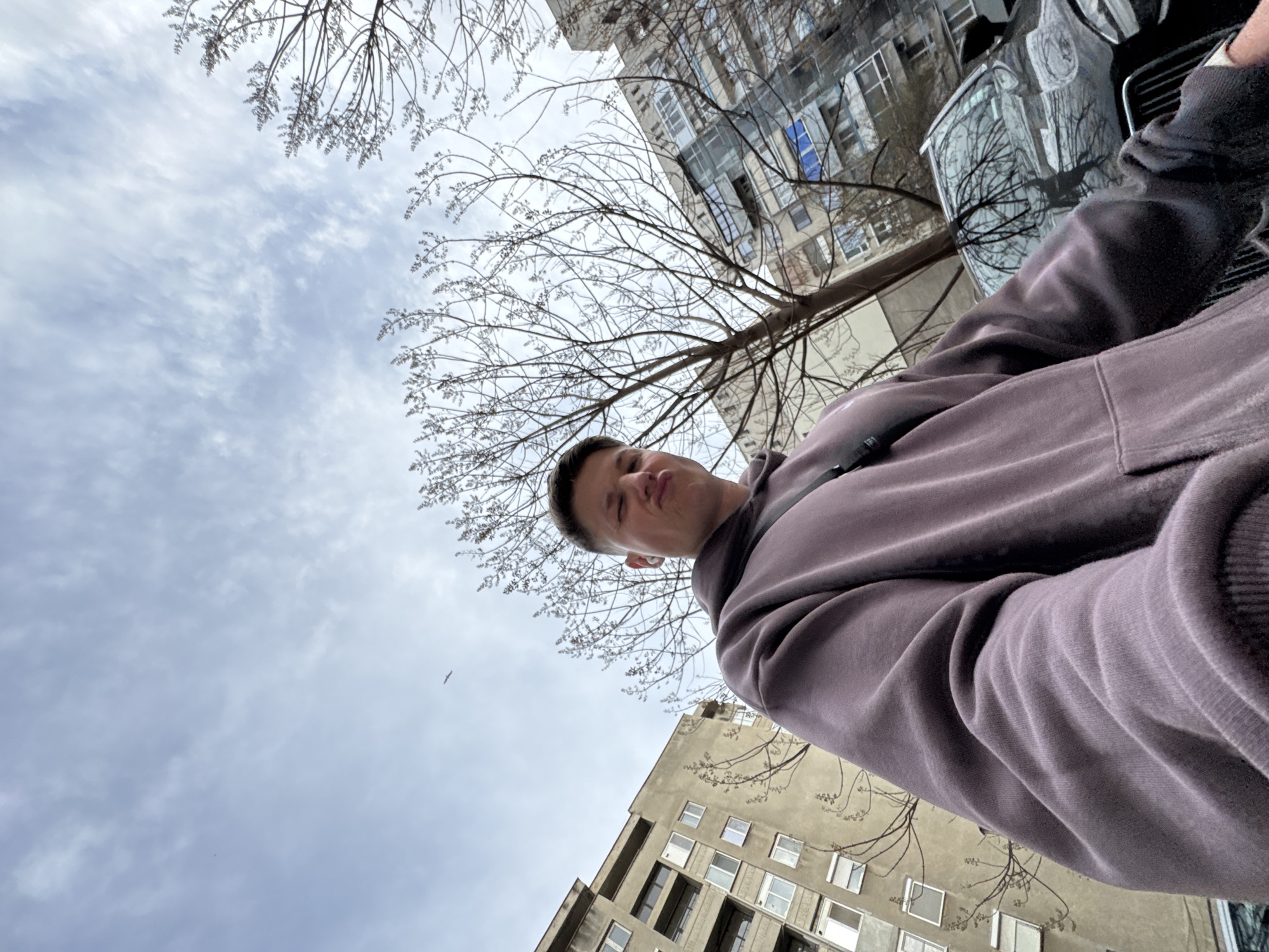 A low-angle selfie of a fair-skinned young man wearing a plum-colored hooded sweatshirt, taken on a city street. The man is positioned in the center of the frame. Behind him, the sky is overcast with a scattering of clouds. In the background, trees with bare branches reach upward toward the sky. Several multi-story buildings with apartments are visible. A car is partially visible in the upper-right corner. The location appears to be in an urban area of Plovdiv, Bulgaria.