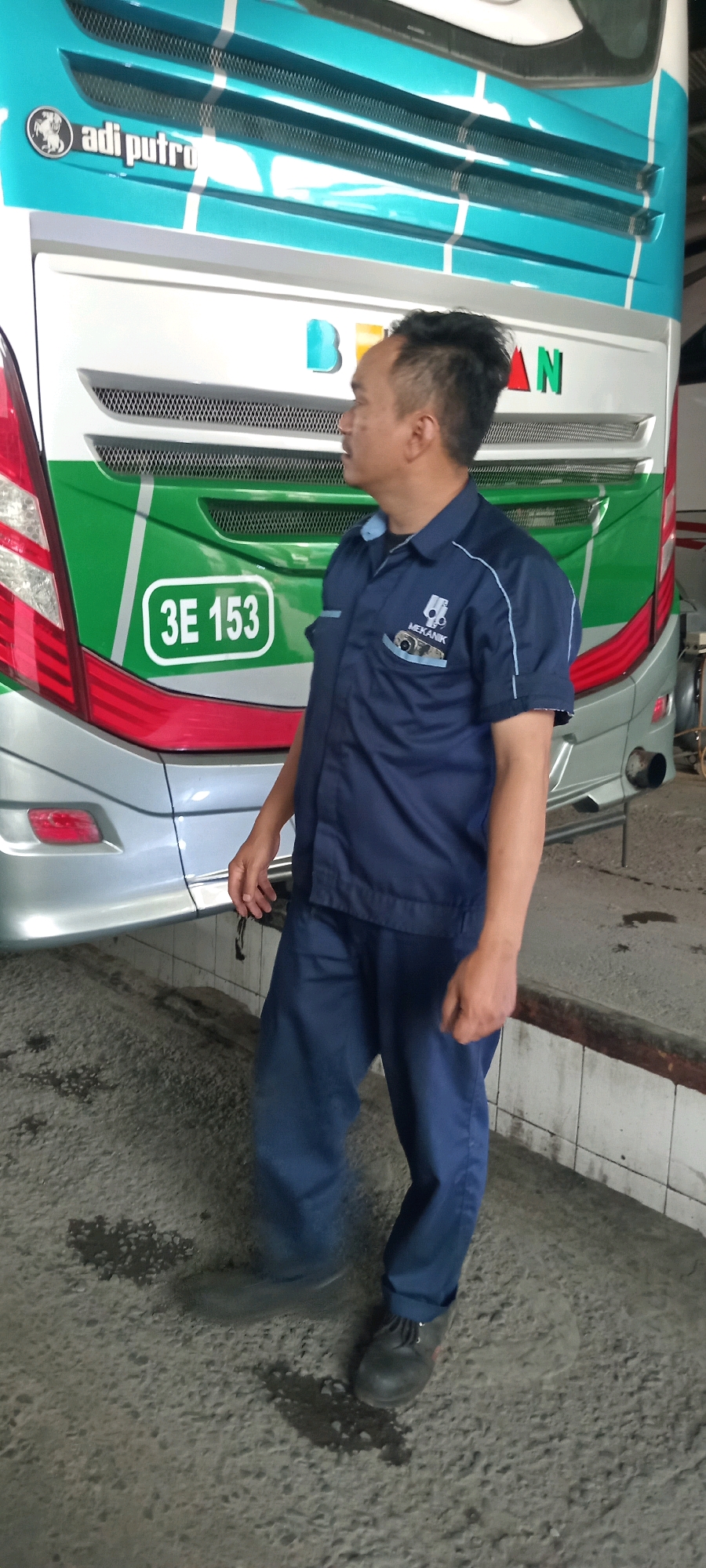 The image shows a man wearing a dark blue mechanic's uniform standing in front of the back of a turquoise and white bus. The bus has the "adi putro" logo on the upper rear section, and a license plate that reads "3E 153" in a white oval. The man is looking to his right, with his hands by his sides. The ground is concrete, with some dark patches that appear to be oil or water stains. The environment suggests an outdoor area, possibly a bus depot or maintenance yard, which could be located within the city of Tasikmalaya, Indonesia, given the commonality of such bus companies in the region. The lighting appears to be natural daylight, with some shadows cast on the ground.