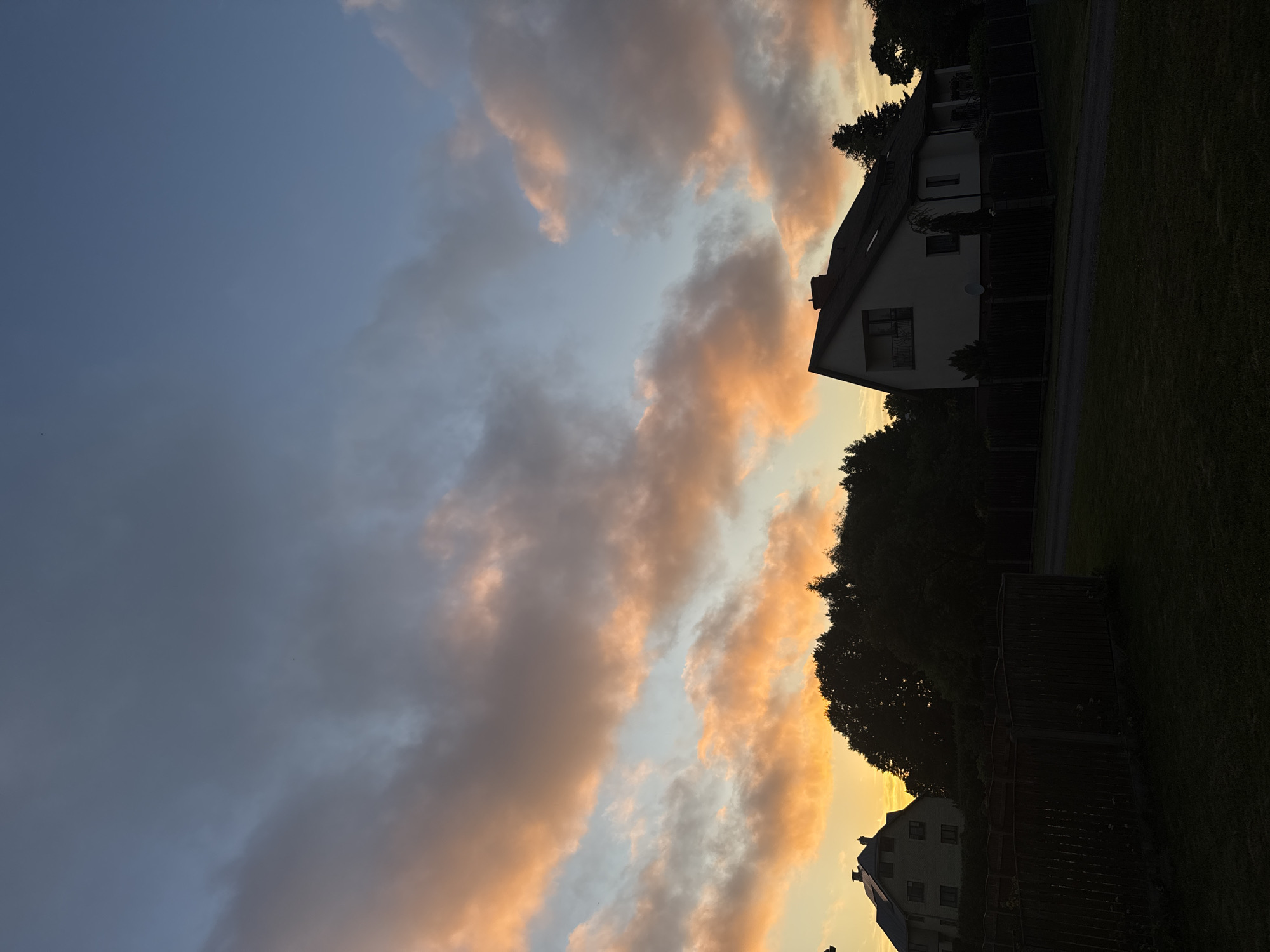 This image depicts a twilight scene in Carnikava, Latvia. The sky is a gradient of soft blues and grays, with streaks of vibrant orange and pink clouds illuminated by the setting sun. Silhouetted against the colorful sky are the dark shapes of trees and two houses. In the foreground, a dark lawn and a wooden fence create a sense of depth. One house, on the right, is a modern white structure with dark trim and a prominent roofline. The other house, on the left, appears to be larger and older, with multiple windows visible. The overall atmosphere is serene and tranquil, capturing the beauty of the fading daylight over a residential area.