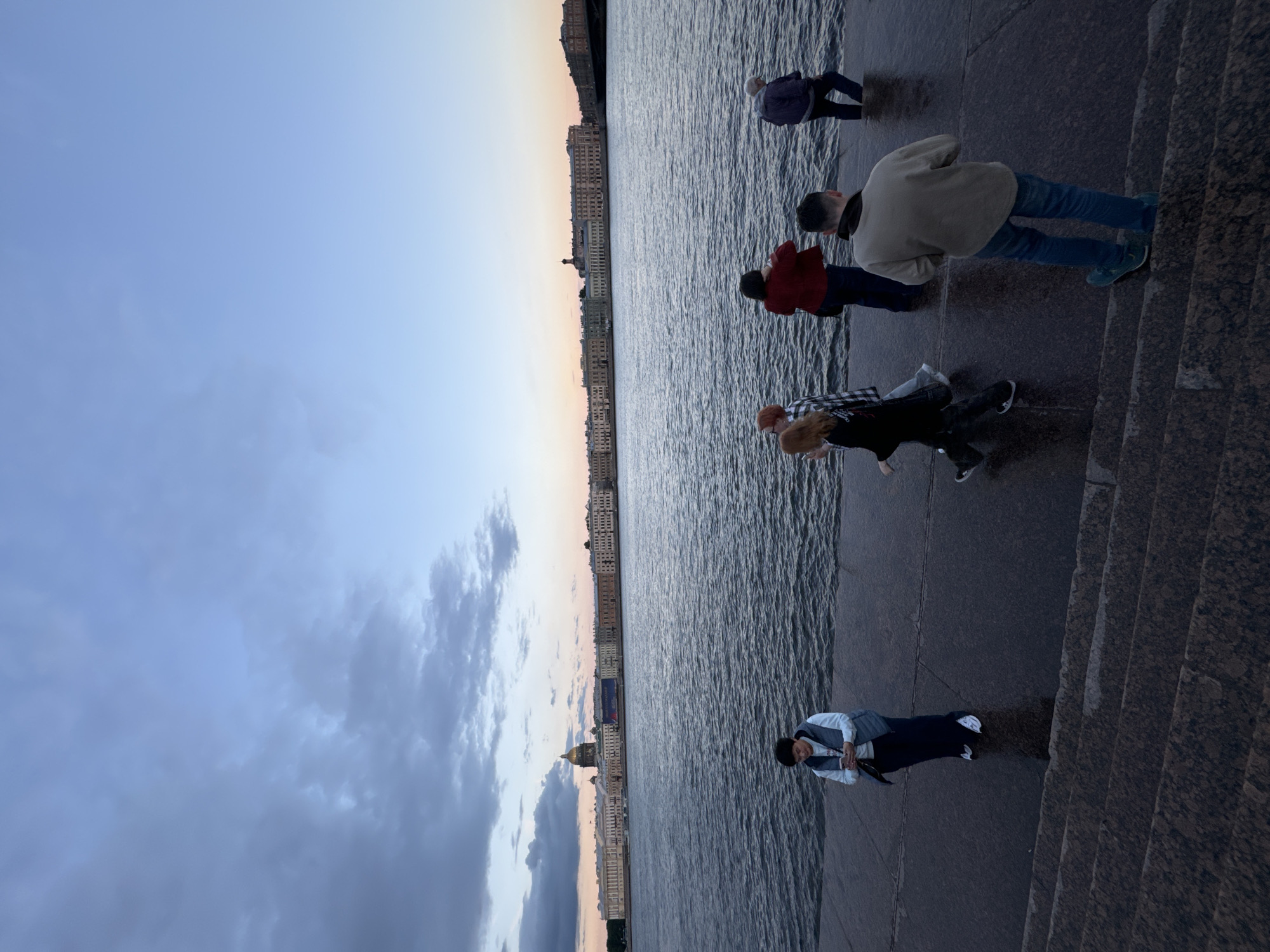 In Saint Petersburg, Russia, at dusk, a group of individuals stands along the Neva River embankment. The city's skyline is visible across the water, featuring a prominent dome building, likely St. Isaac's Cathedral, and a long row of classical architecture. The sky is a gradient of blue and pink hues, with scattered clouds.
On the embankment, which appears to be constructed of granite, several people are walking or standing. One person in the foreground, facing away from the camera, is wearing a beige sweatshirt and blue jeans, with teal sneakers. To their left, a person in a dark jacket and blue pants walks towards the water. Further left, a person wearing a light blue vest over a white top and dark pants stands looking at their phone. To the right, a person in a dark jacket and blue jeans is partially in the water. Other individuals, including two women, are also present, some looking towards the city across the river. The water of the Neva River is rippling, reflecting the twilight sky. The scene conveys a sense of quiet evening activity by the river.