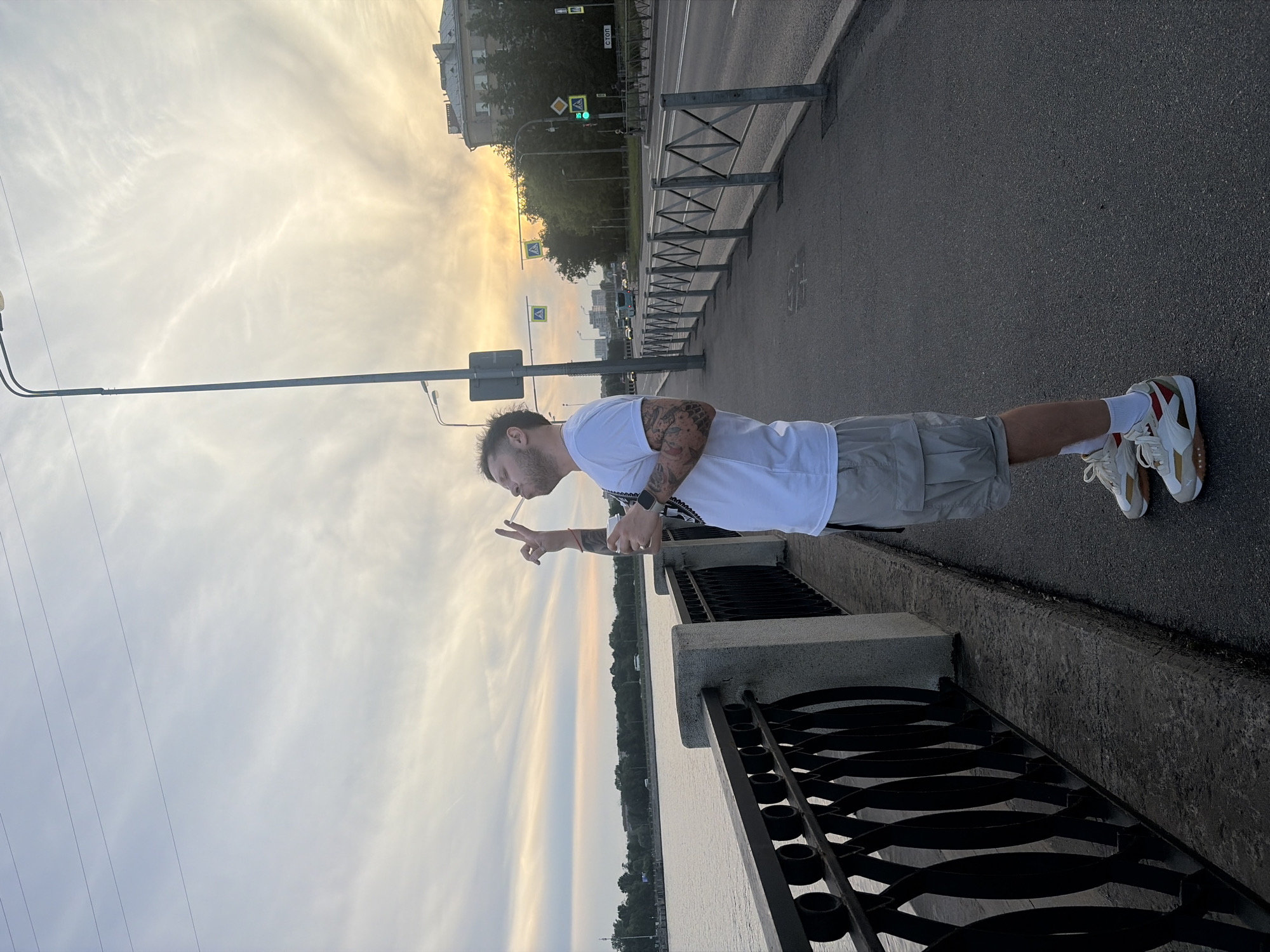 A male individual stands facing left on a pedestrian walkway adjacent to a river embankment in Saint Petersburg, Russia, under a warm sunset or sunrise sky. The man, with short dark hair and a beard, is dressed in a white short-sleeved t-shirt, light cargo shorts, and white sneakers with red and gold accents. Visible tattoos adorn both of his forearms, and a dark smartwatch is on his left wrist. He holds a white cup in his left hand and a translucent plastic straw or bubble wand with his right hand, positioning it near his mouth while extending his index and middle fingers upwards in a "V" shape. He looks slightly upwards and to his left.
To his left, a decorative black metal railing with circular motifs tops a concrete embankment wall, overlooking a wide river or canal. The far bank of the water body is lined with trees and distant buildings. To the man's right, a multi-lane asphalt road runs parallel to the embankment, separated from the walkway by a lower metal barrier. Along the road, several streetlights, traffic signals (one displaying green), and pedestrian crossing signs are visible, including a Russian "СТОП" sign. Vehicles and a bus can be seen on the road. Further right and behind the road, a multi-story building with a distinctive, complex roofline stands amidst trees. The sky transitions from light blue at the top to a warm orange and yellow hue near the horizon, streaked with wispy clouds and crossed by faint power lines.