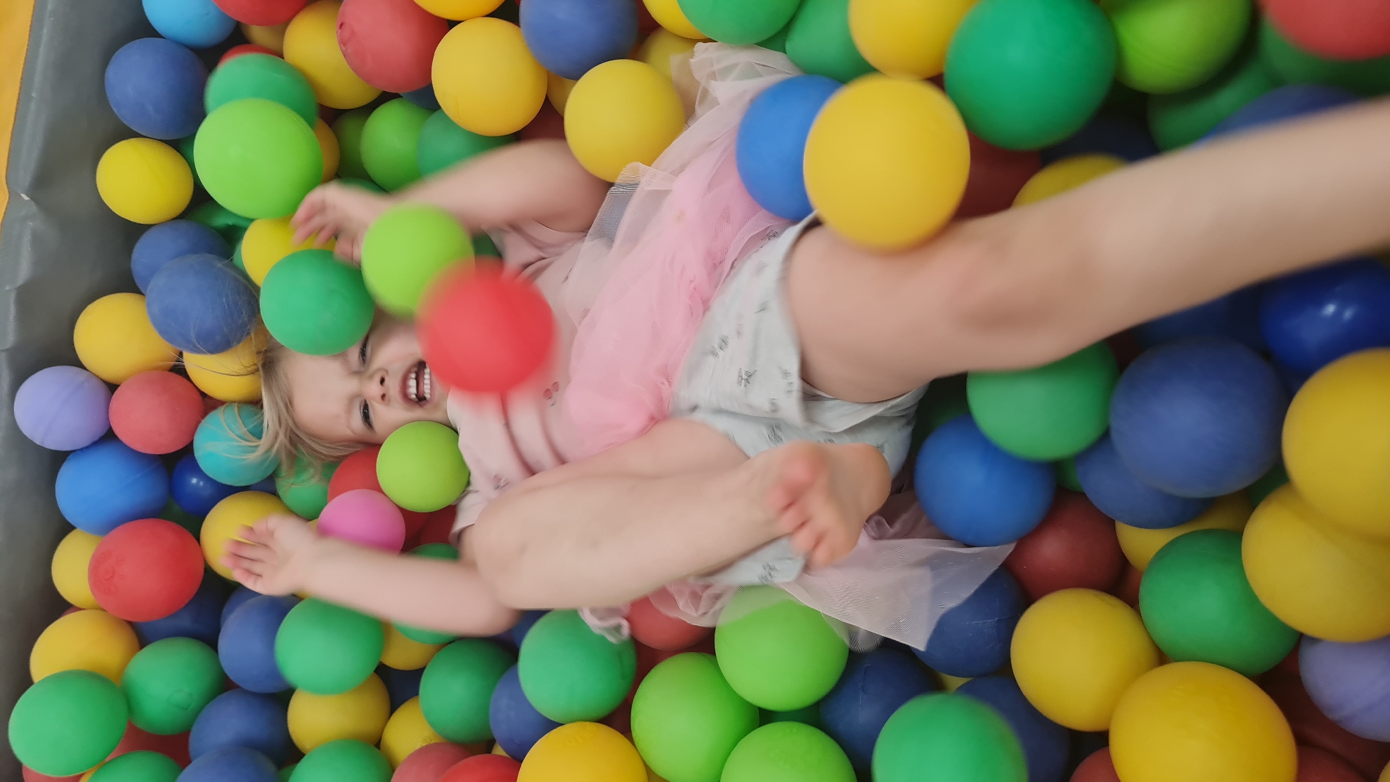A child is in a ball pit filled with yellow, green, blue, and red plastic balls. The child is wearing a pink shirt and shorts and appears to be falling backward, with legs extended. The background is filled with more of the colorful plastic balls. The location is Lachish Regional Council, Israel.