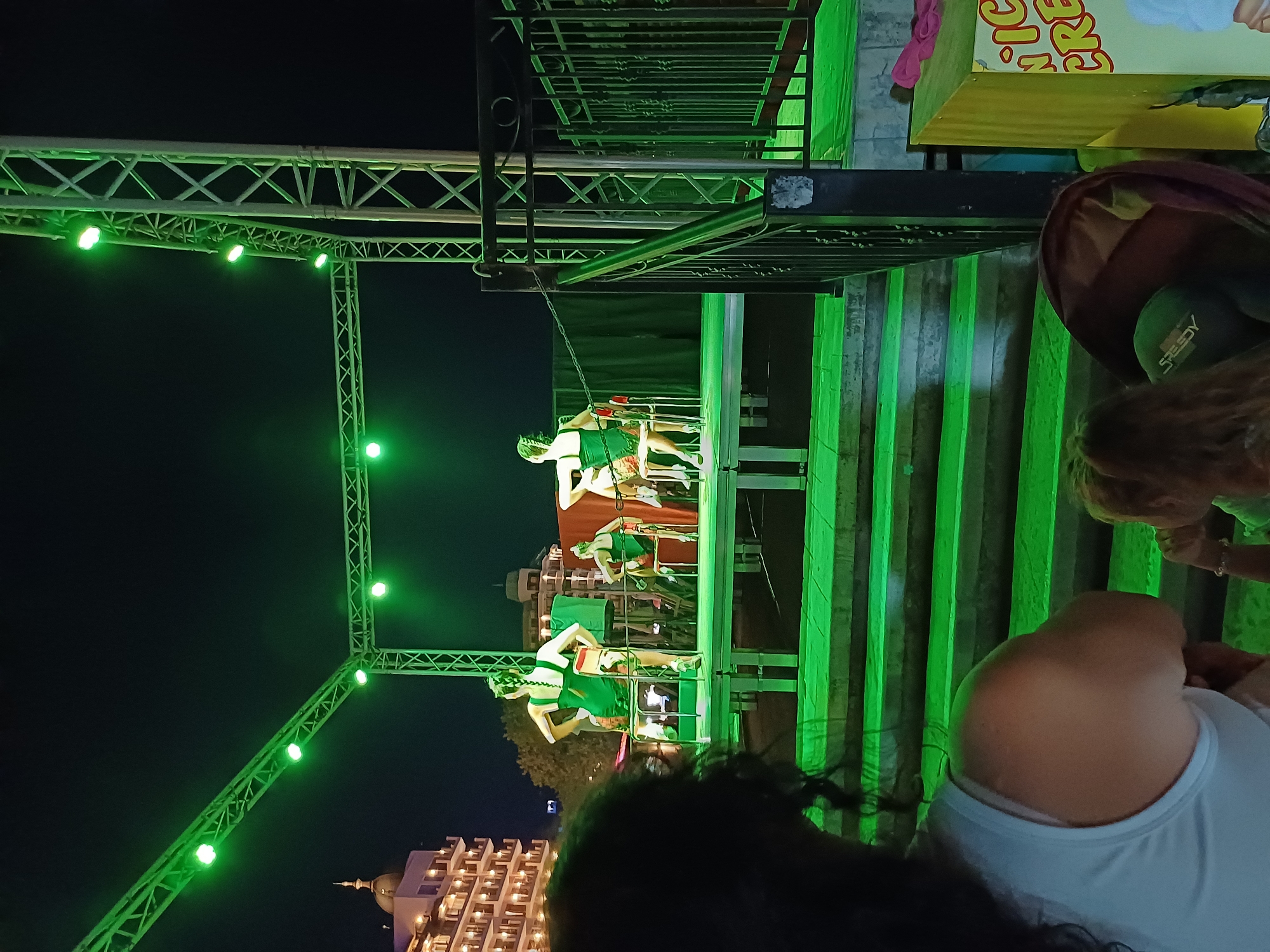 In Varna, Bulgaria, an outdoor performance is taking place at night, illuminated by green stage lights. On a raised platform, three women dressed in dark green leotards and colorful skirts are seated on chairs. Two of the women are facing away from the camera, while the third is partially visible from the side. The stage is supported by metal scaffolding and backed by a dark curtain. In the foreground, the back of a woman's head and shoulder is visible, suggesting she is part of the audience. A child's head is also visible to her right. Behind the stage, a multi-story building with illuminated windows is visible. To the right of the scene, a yellow kiosk with the partial text "ICE CRE" is present. The overall impression is of a lively evening entertainment event.
