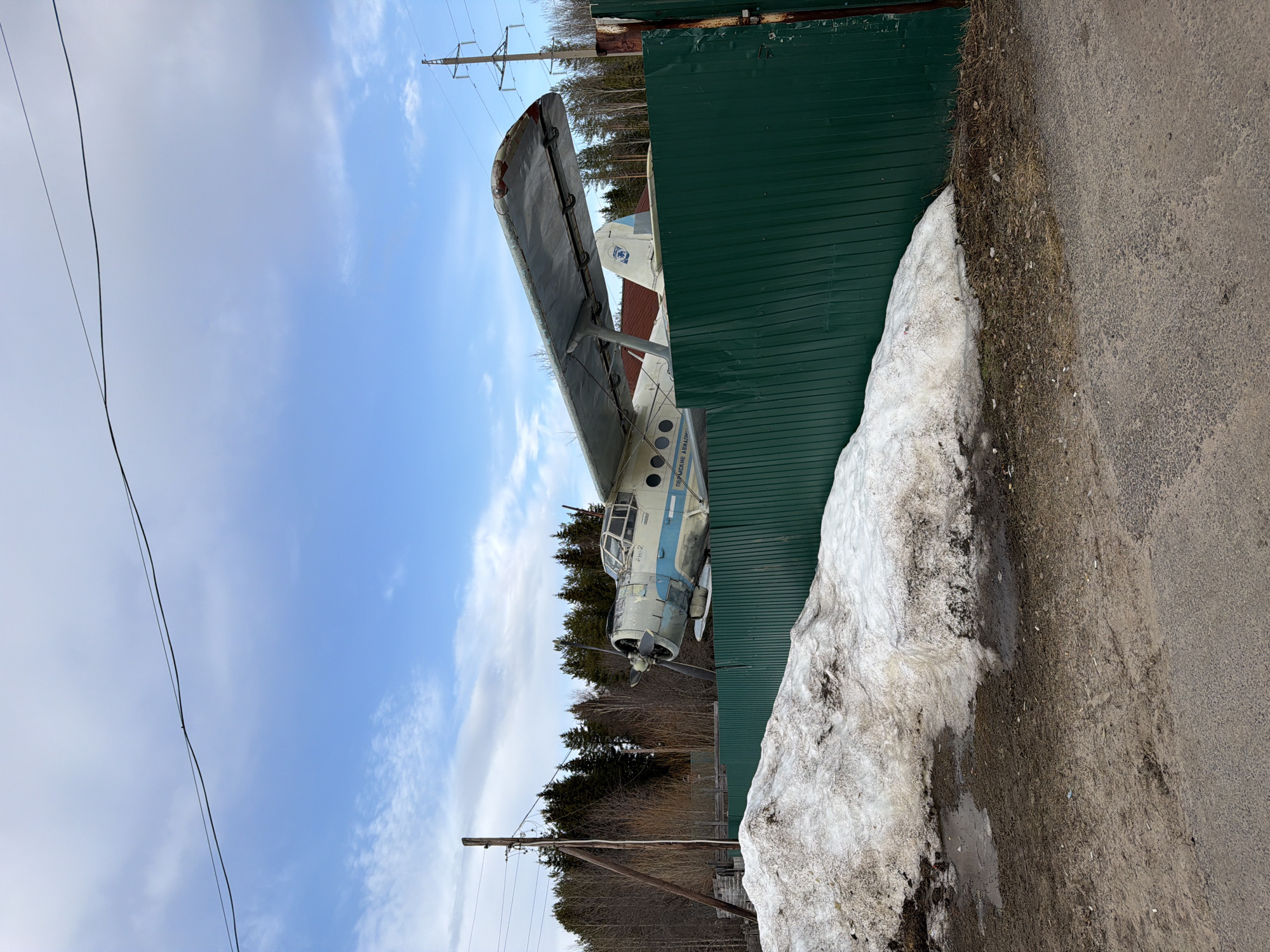 The image shows an Antonov An-2 biplane situated in what appears to be an area near Жуланово, Russia. The aircraft is partially obscured by a green corrugated metal fence. There's a snow pile alongside the fence and the edge of a paved road is visible. Power lines and utility poles can be seen against the blue sky with some clouds. The surrounding environment includes trees, suggesting a location on the edge of a wooded area. The biplane is painted in a light blue and white color scheme, with some visible text in Russian.